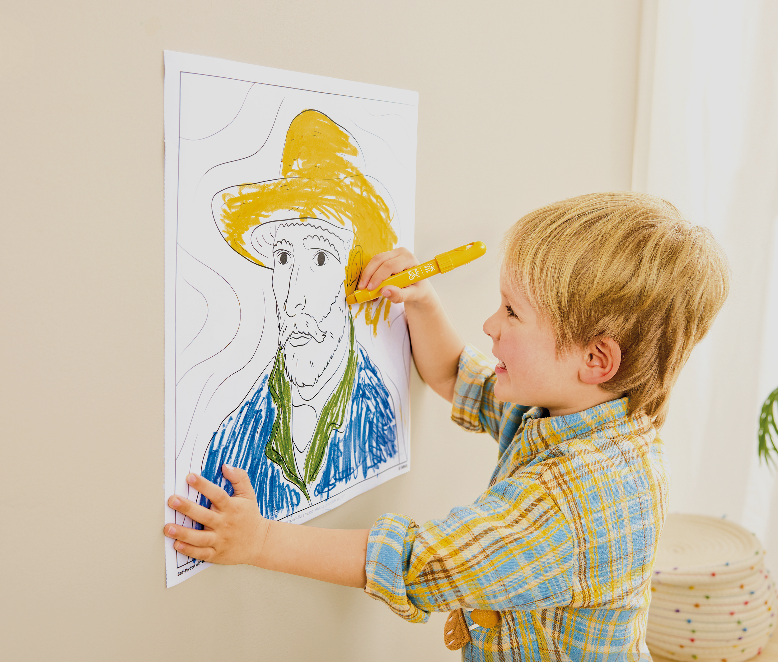 A young child with blonde hair, wearing a yellow and blue plaid shirt, is pasting a yellow and green crayon drawing of a face with a hat onto a wall. The drawing depicts an older man with a beard and a large hat, and the child is holding a yellow cra