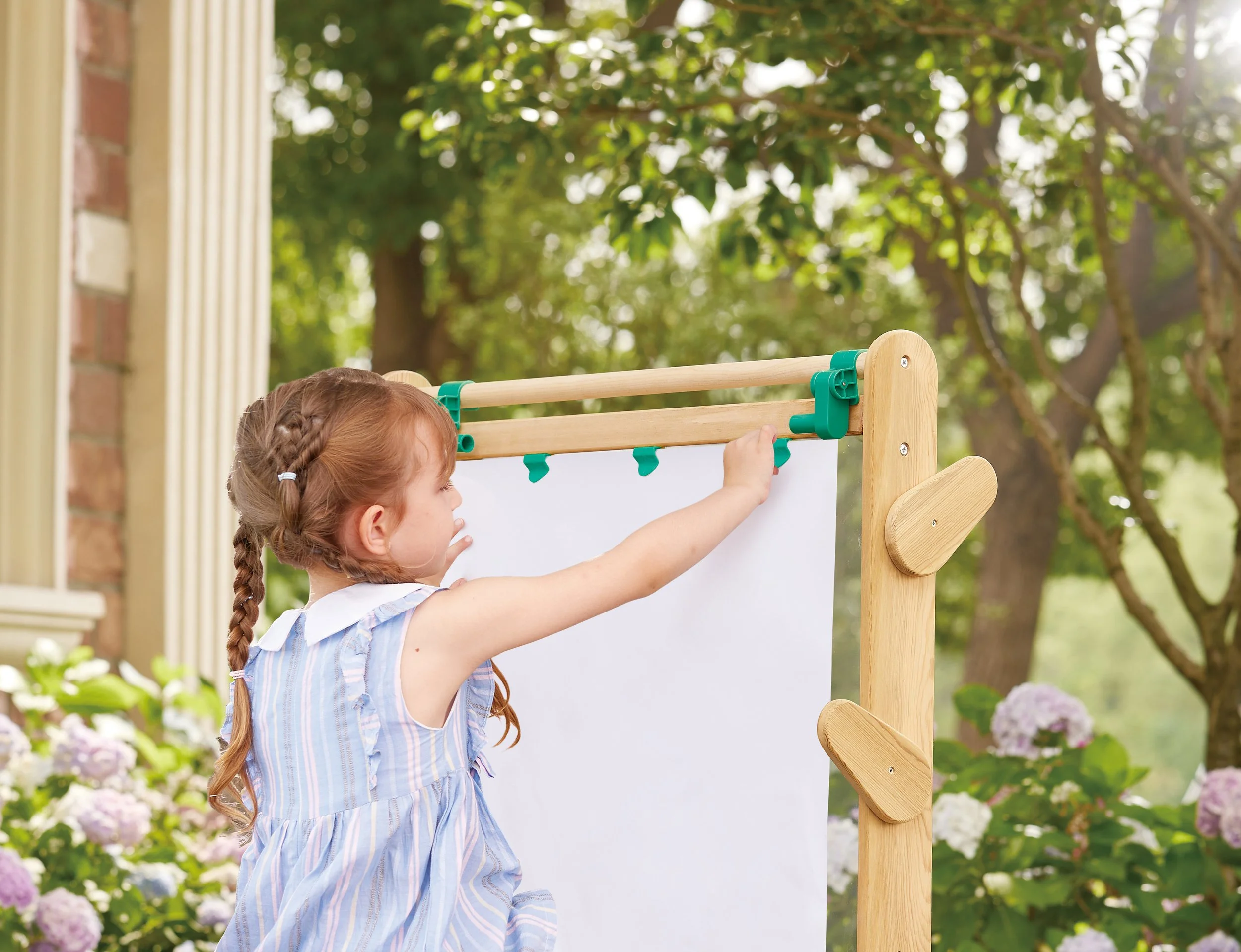 A little girl with her hair braided in a blue and white dress hanging a white sheet on the wooden outdoor Hape board in a garden with green trees and hydrangea flowers.