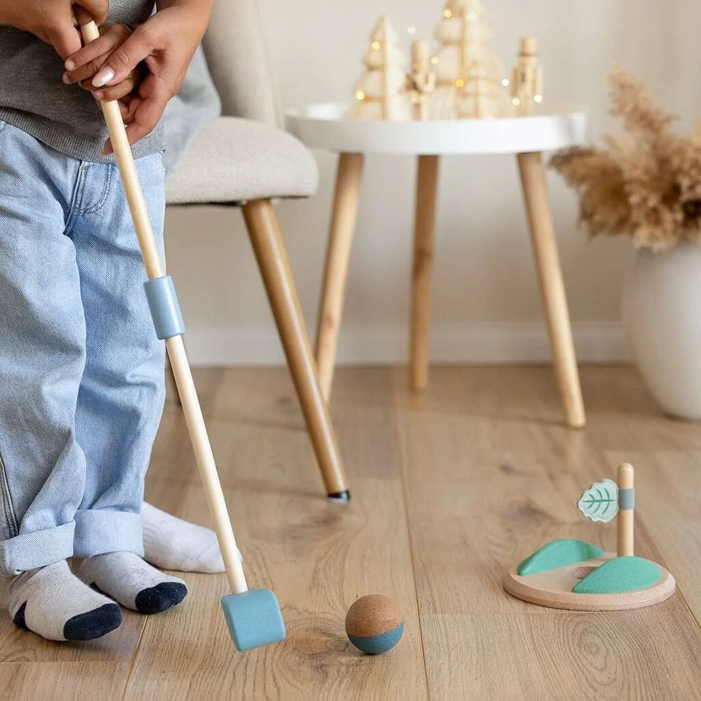 Child in gray sweater and light blue jeans practicing golf indoors with a toy golf set, including a club and ball, on a wooden floor in a cozy living room. By Korko.