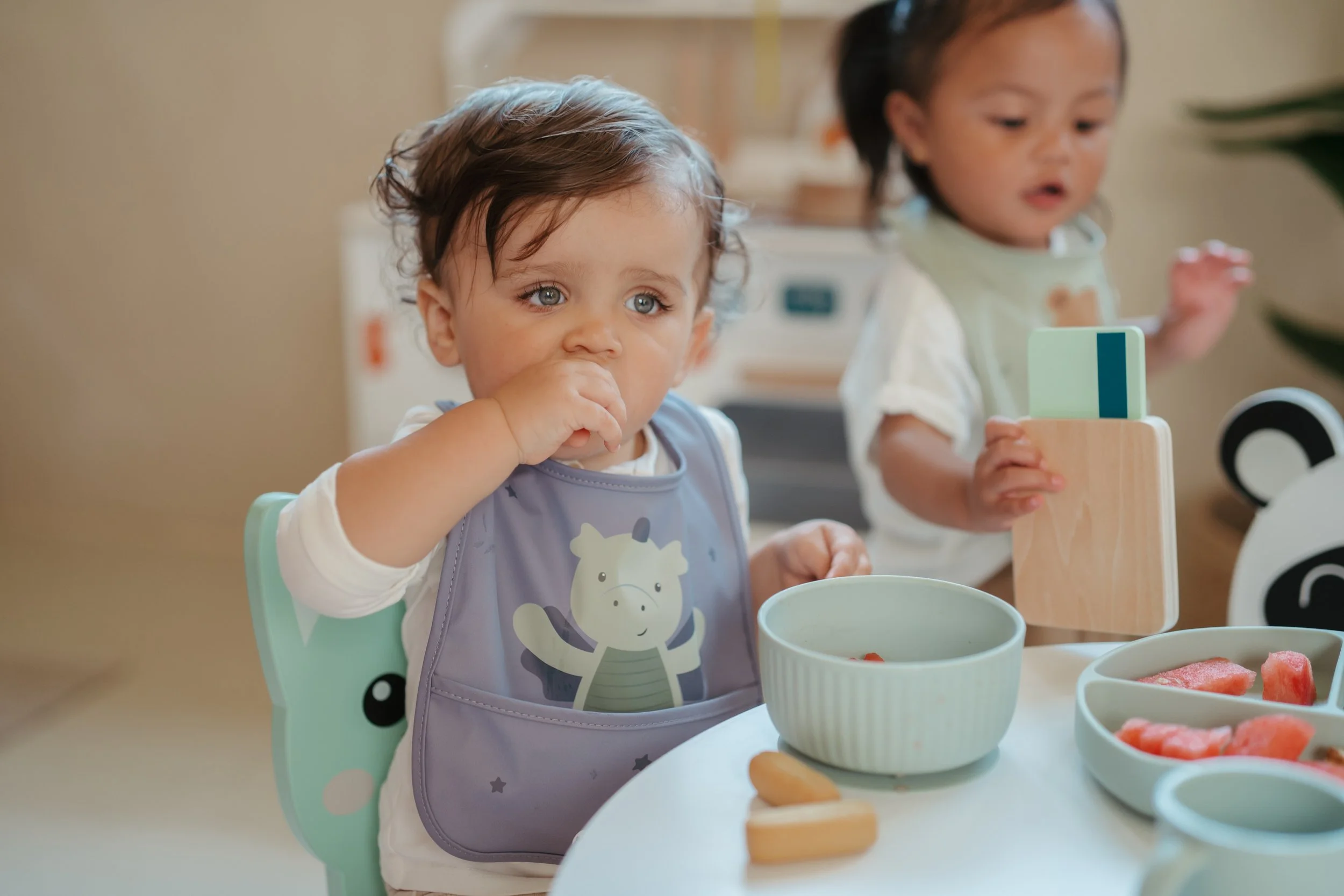 Two young children sitting at a table with bowls of food, one child has a purple bib with a zebra on it and the other is holding wooden utensils.