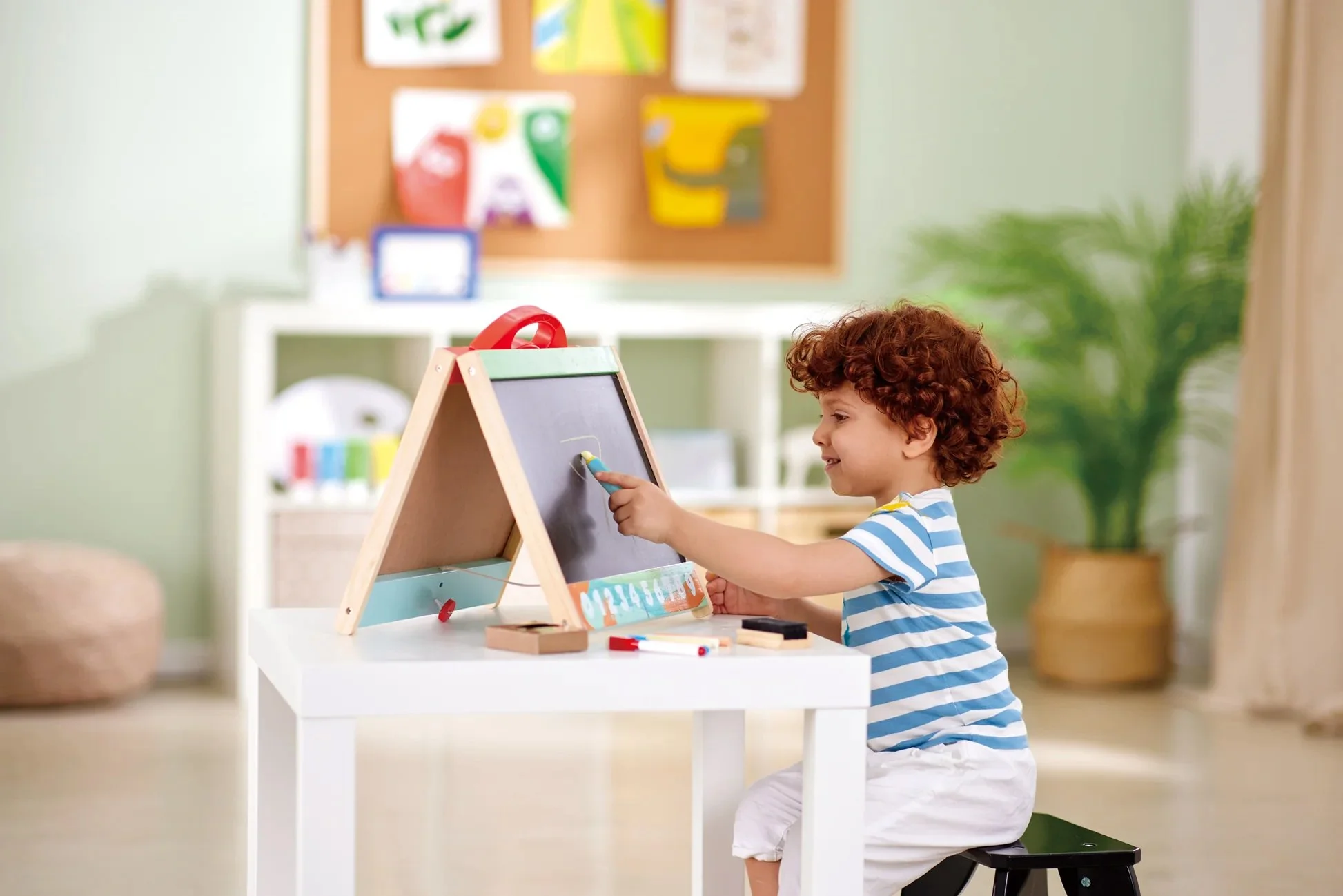 Young boy with curly hair sitting on a small black stool, playing with the Hape chalkboard in a bright, decorated room.