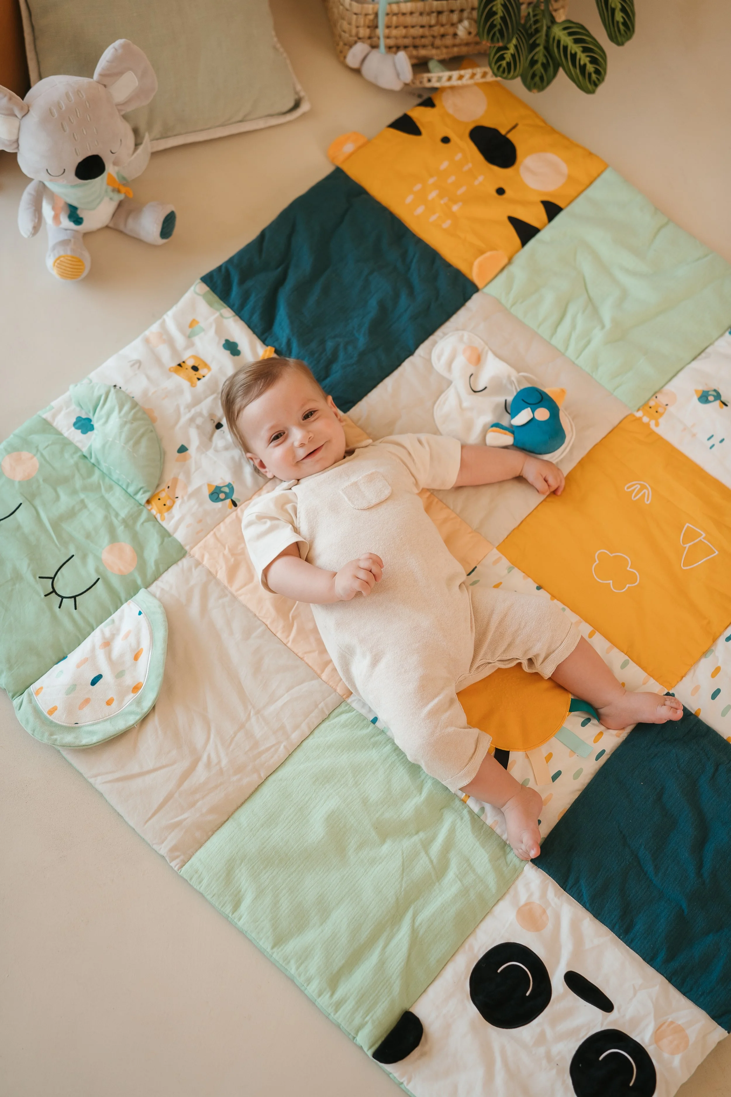 Young child lying on a colorful patchwork quilt with animal and nature prints, smiling. Nearby are plush toys and a stuffed animal with a mask. Decor includes a plant and a basket.