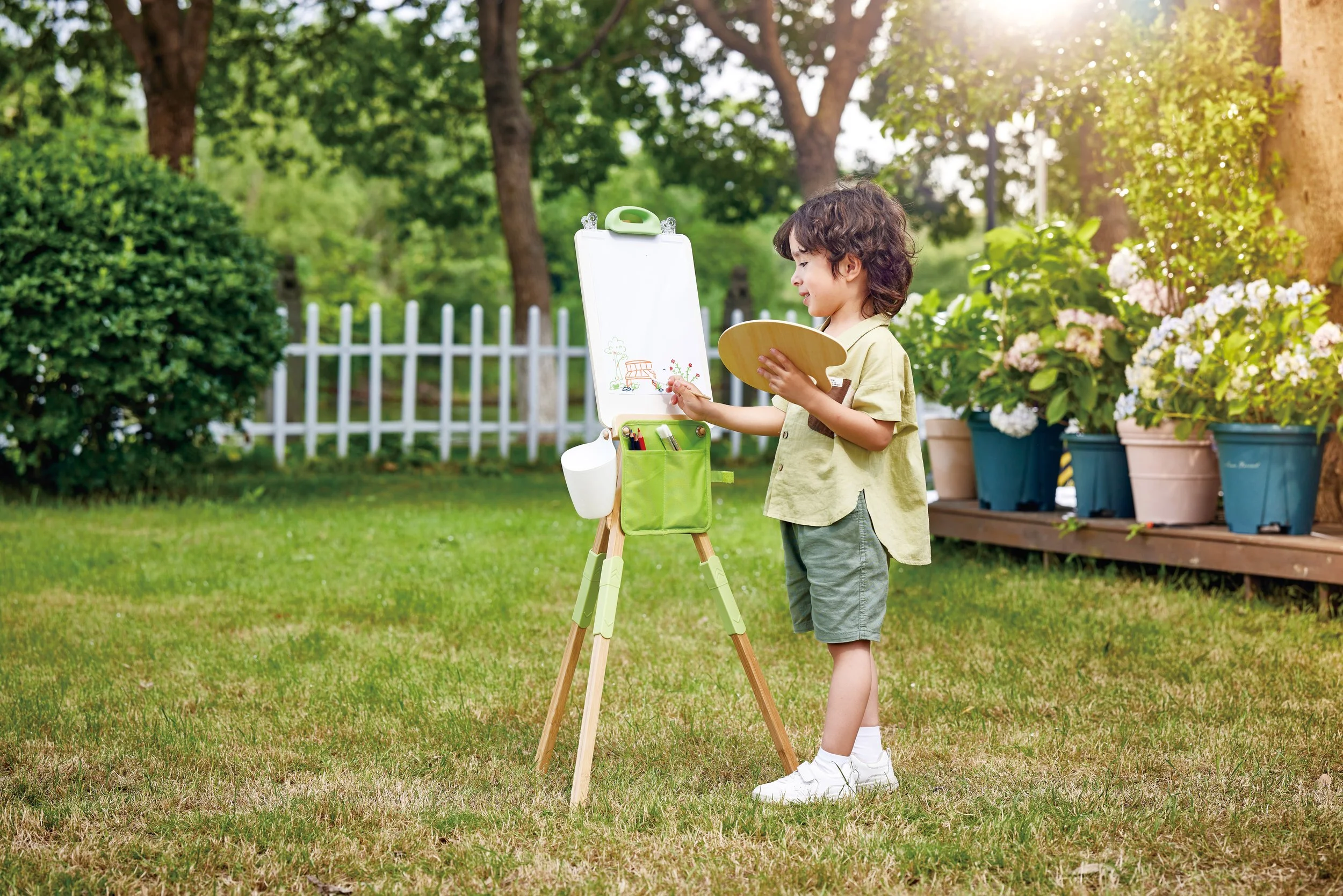 A young boy painting on the outdoor bamboo easel by Hape in a garden with trees and potted plants.