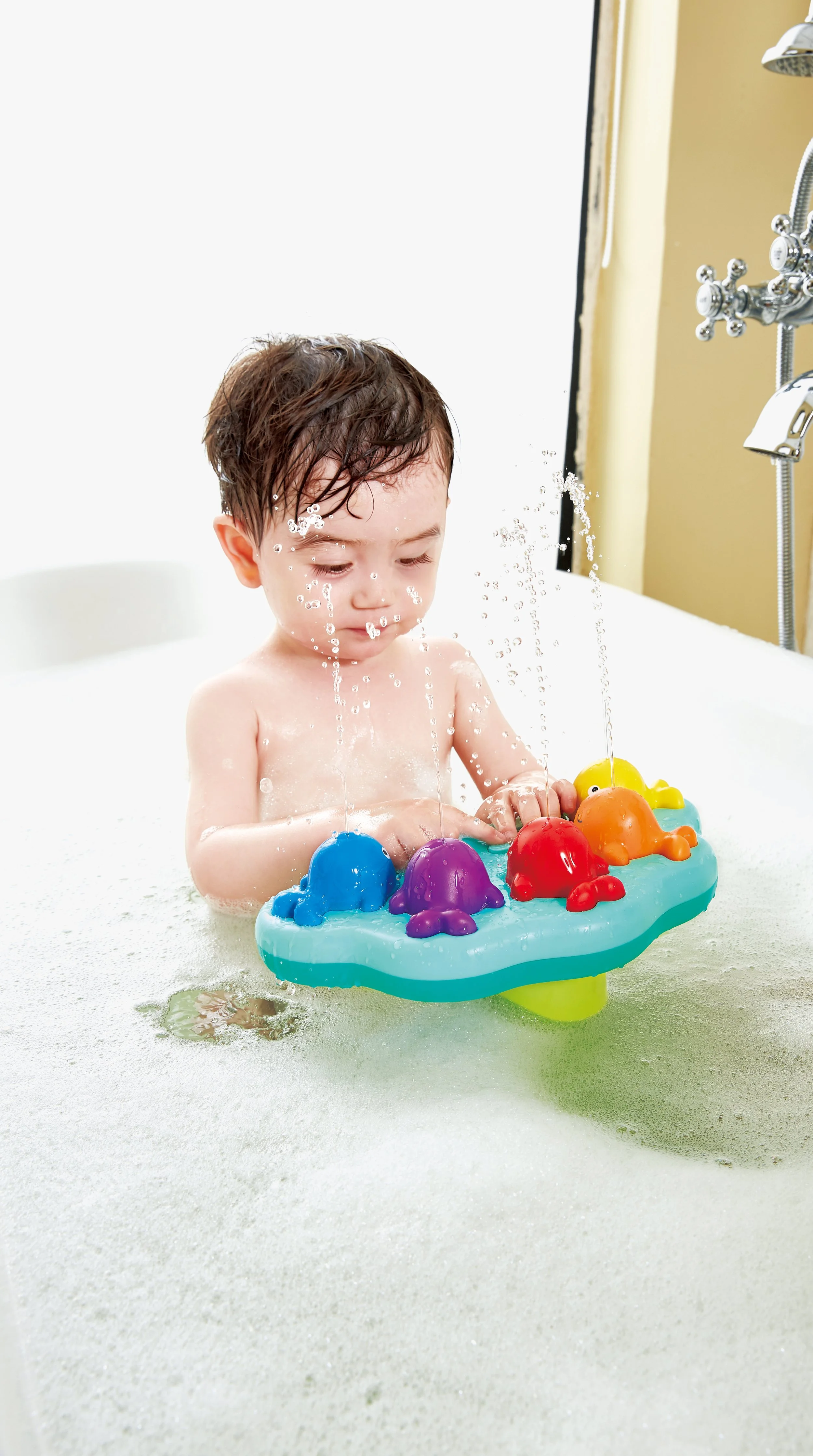 A young child playing in a bathtub with "Musical whale fountain" by Hape, surrounded by soap bubbles, with water splashing.