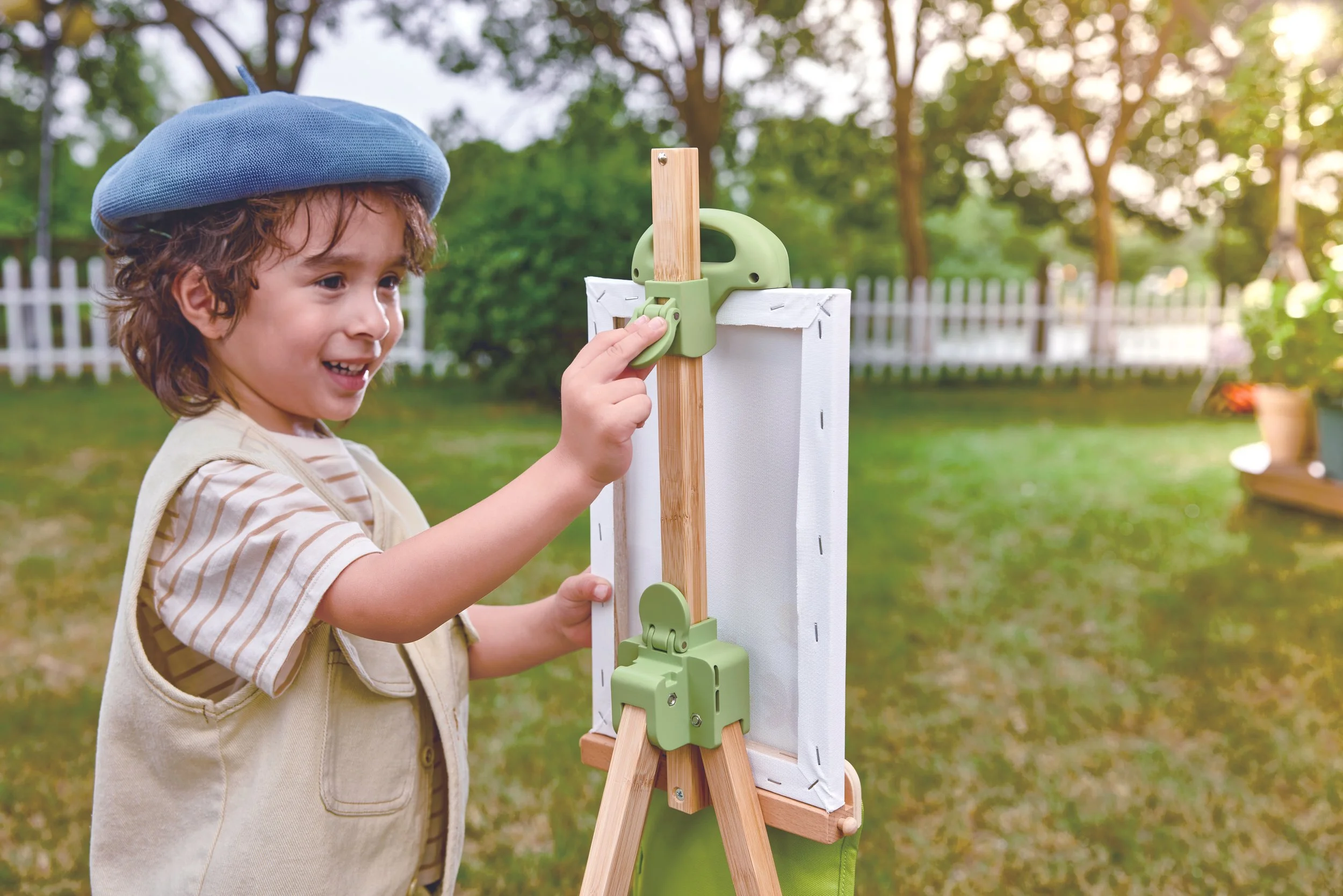 A young boy with curly hair, wearing a blue beret and a beige vest over a striped shirt, playing with the bamboo easel by Hape outside in a grassy backyard with a white picket fence and trees, during sunset.