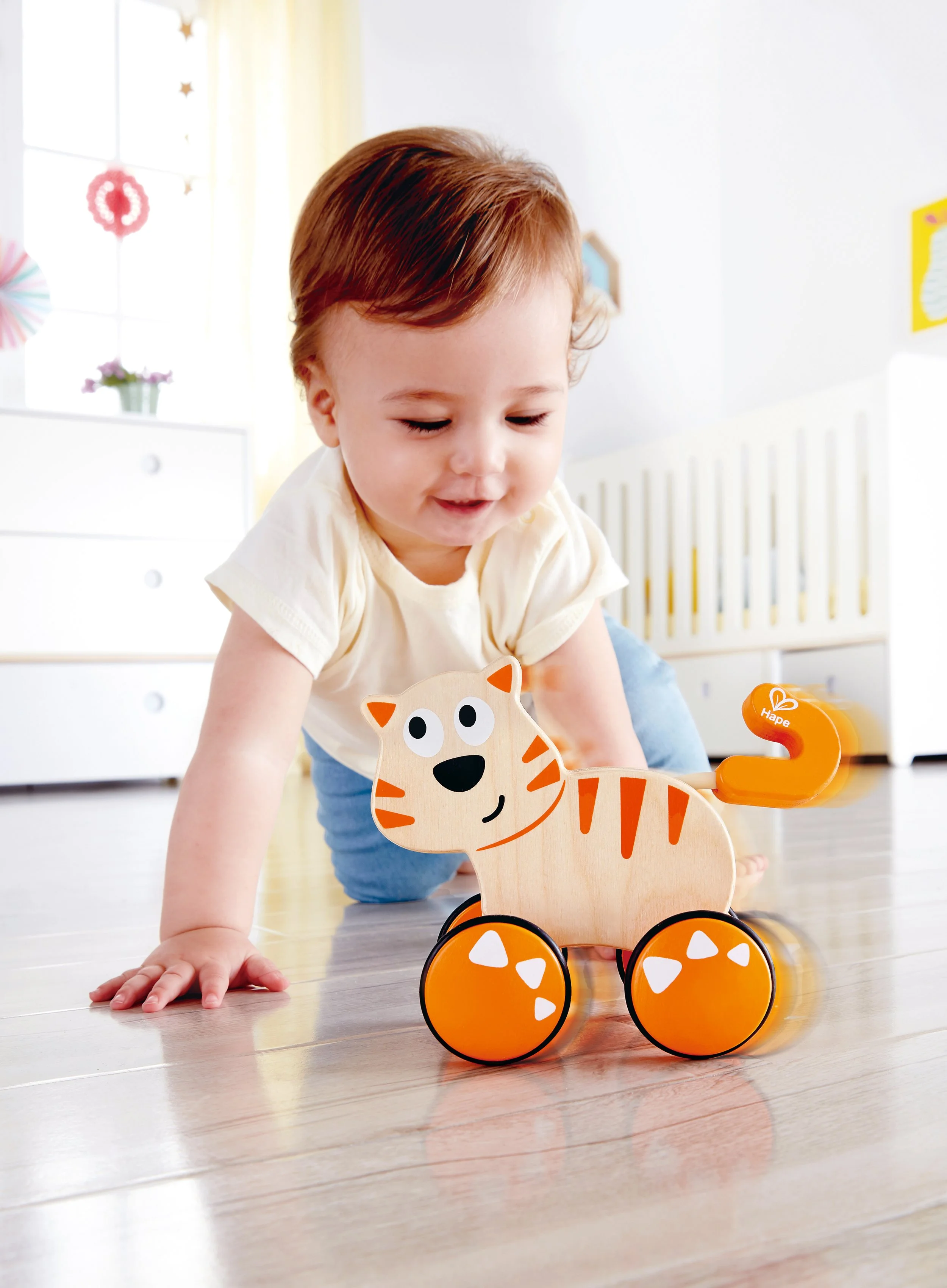 A young child with brown hair is crawling on a light wooden floor, playing with a wooden cat-shaped toy on wheels in a bright, colorful room. Developed by Hape.