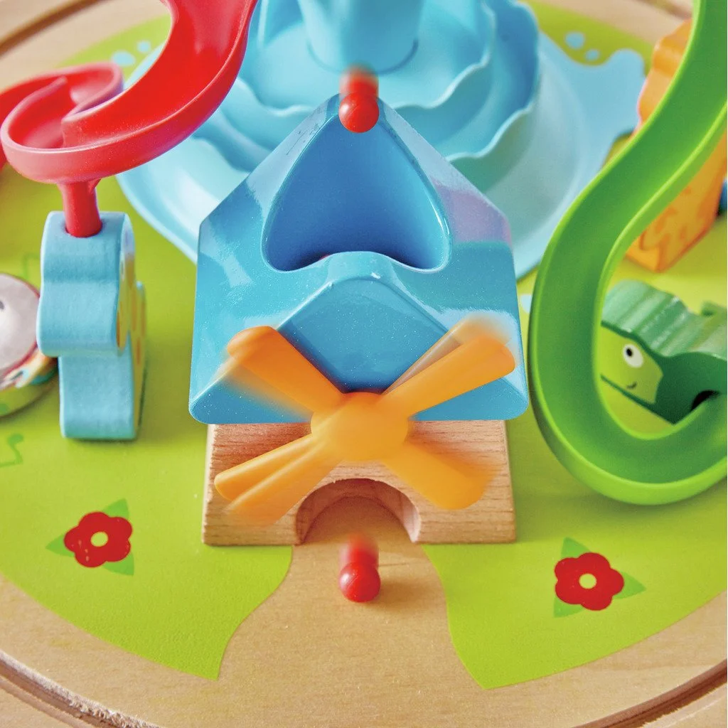 Colorful children's marble run toy with plastic tubes, a roller, and a wooden base, featuring a green snake decoration and small red flower illustrations on a yellow background. Developed by Hape.