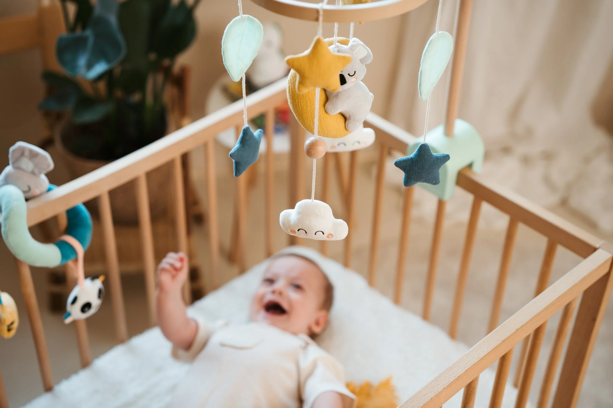 Smiling baby lying in a wooden crib, looking up at hanging felt mobile with stars, clouds, moon, and stuffed animals.