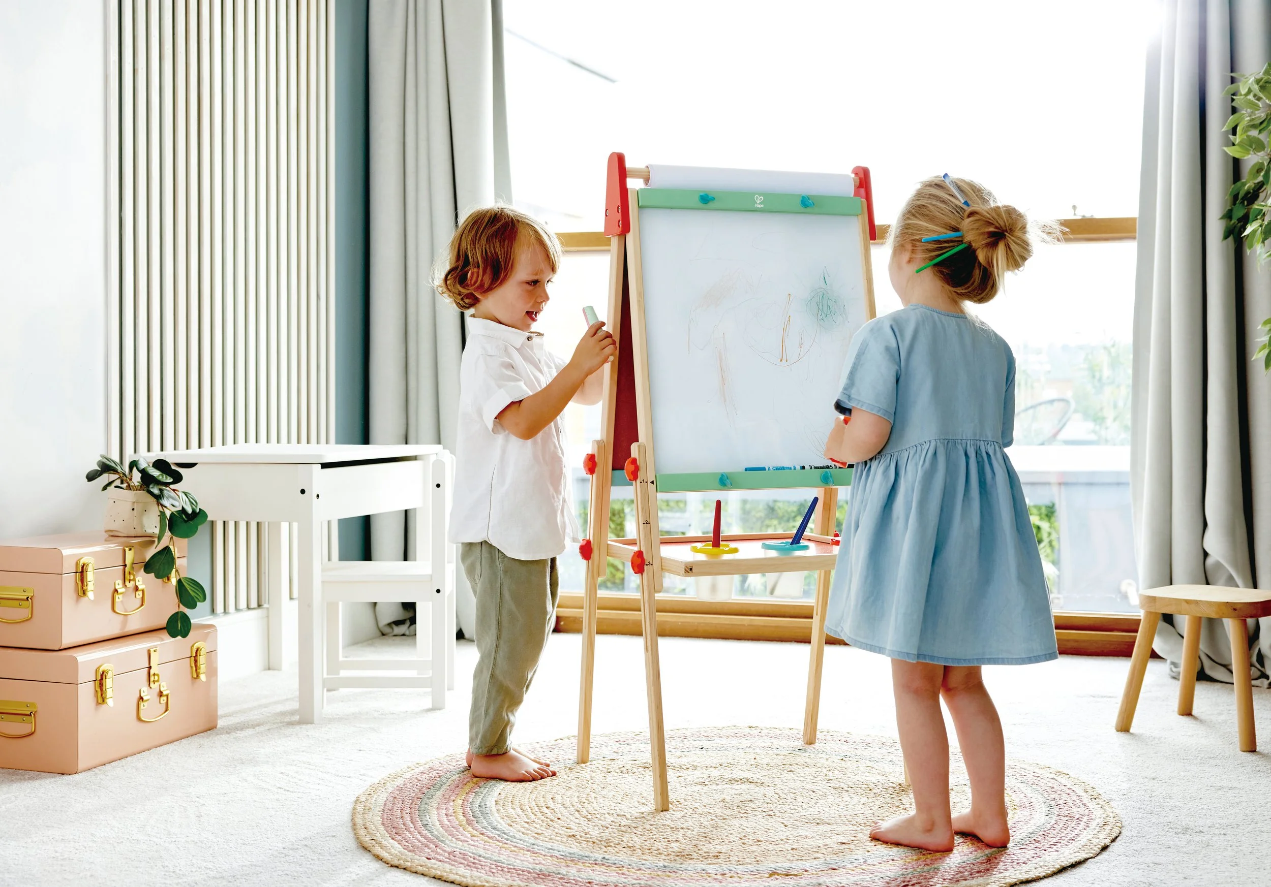 Two young children, a boy and a girl, are drawing on the Hape whiteboard in a bright room with large windows and curtains. The boy, with curly hair, is holding a chalk and smiling. The girl, with her hair in a bun, is standing with her back to the ca