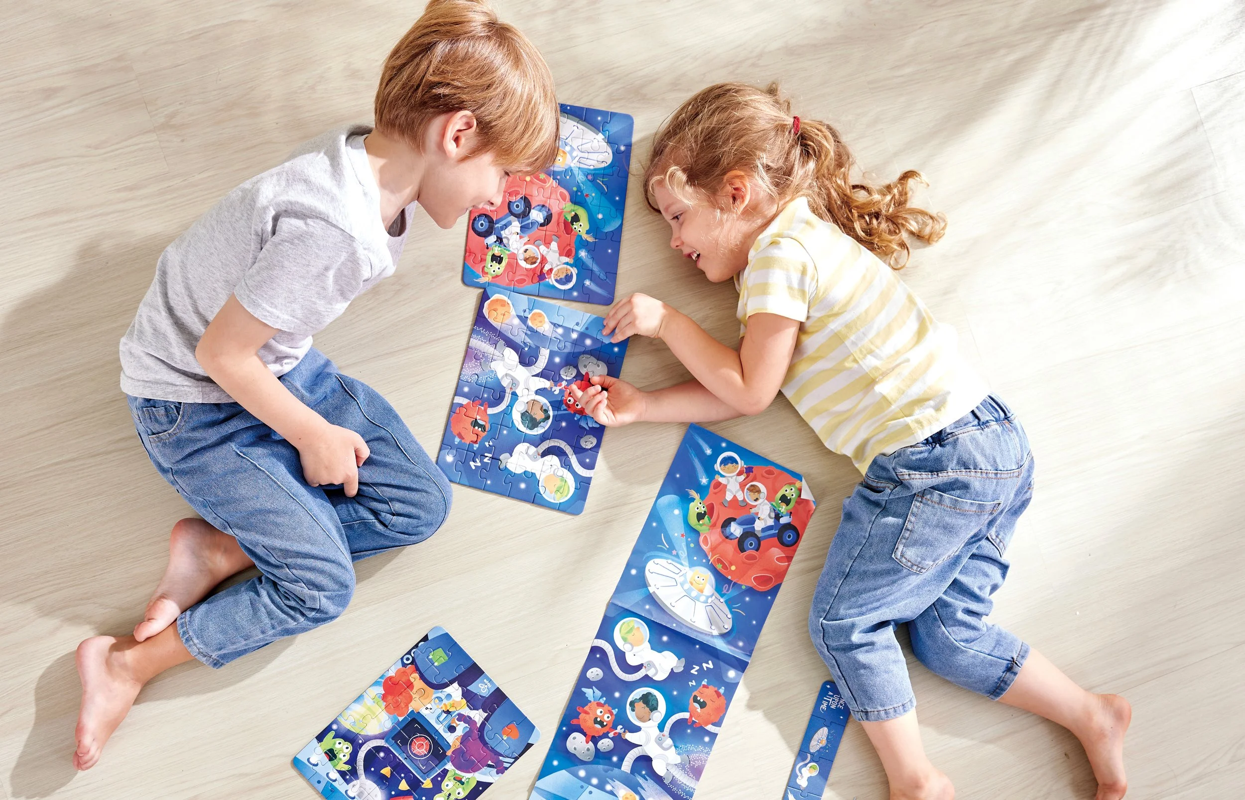 Two children, a boy and a girl, lying on the floor and assembling a space-themed Hape puzzle with images of astronauts, rockets, and planets.