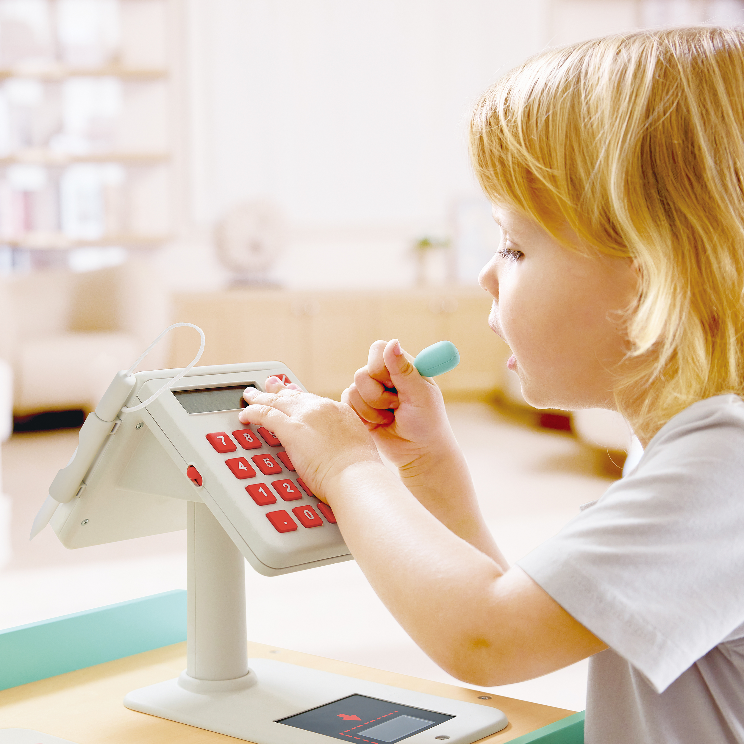 A young girl with blonde hair at a toy cash register, pressing buttons with one hand and holding a toy credit card with the other.