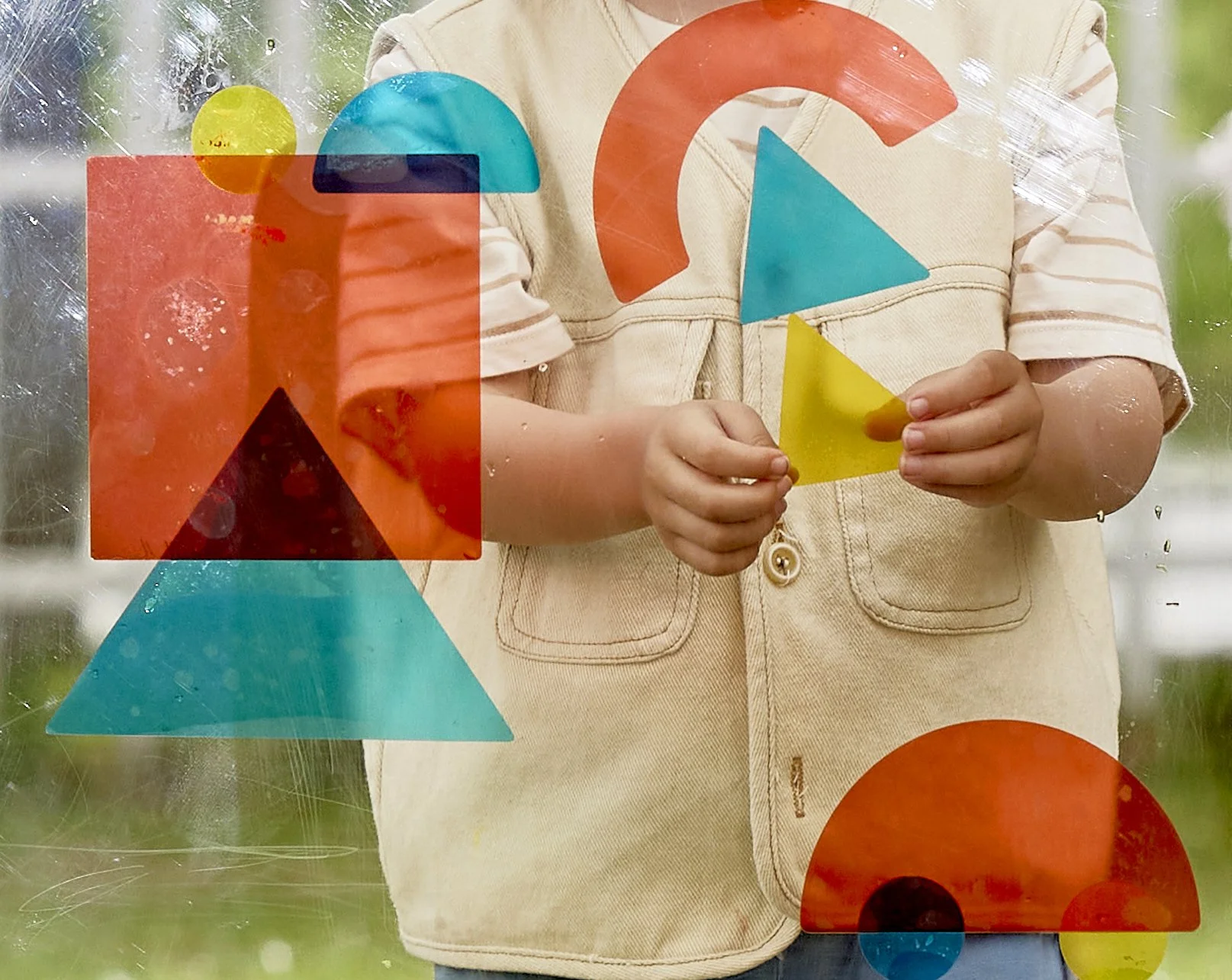Child wearing a beige jacket with striped sleeves, holding colorful geometric shapes (triangle, square, circle) on a glass surface.