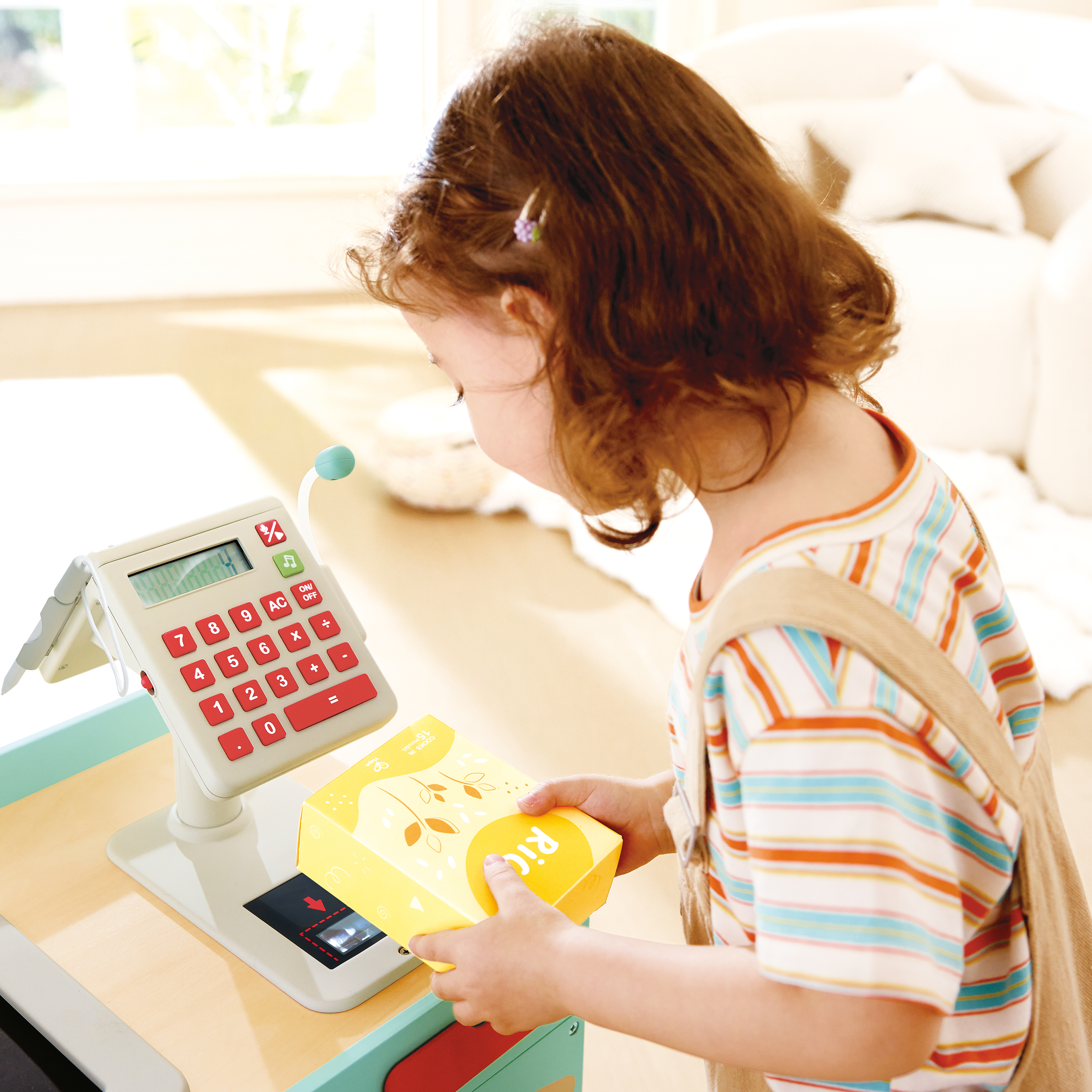 Child with red hair using a toy cash register to scan a yellow box labeled 'RIL' in a sunny, cozy room.