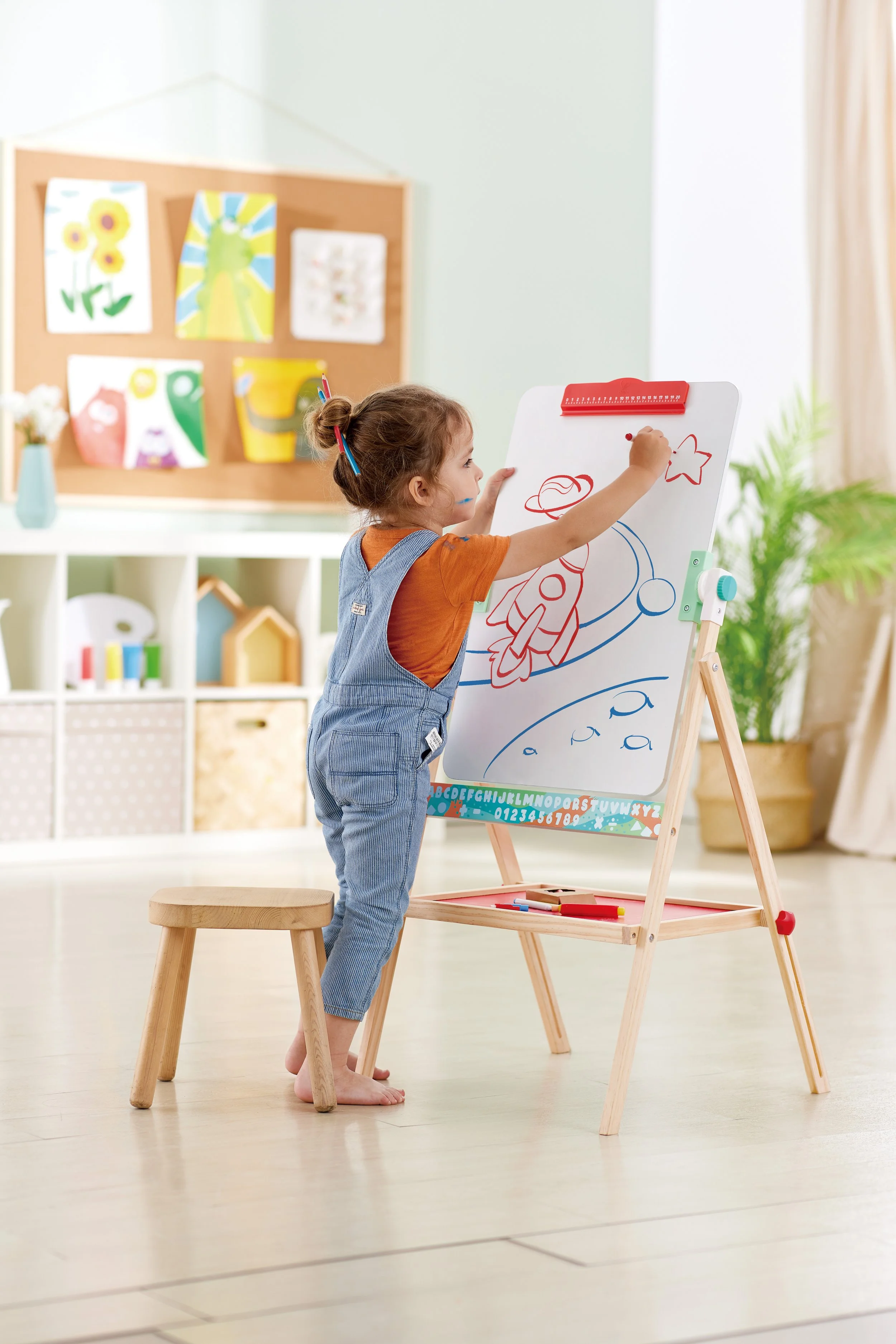 A young girl drawing on the Hape whiteboard with colored markers in a bright, cozy playroom.