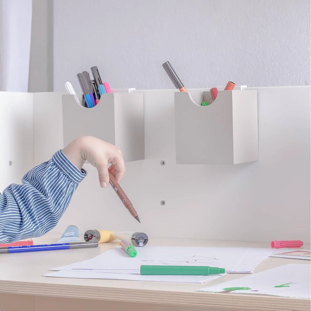 Child's hand with a brown marker over a Colorflex desk with drawing papers, colorful markers, and scattered art supplies, with a white wall and storage bins holding more markers in the background.