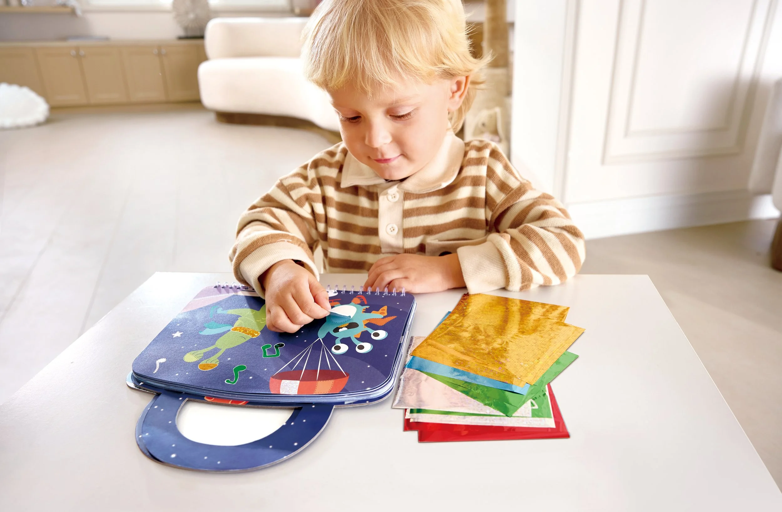A little boy with blond hair sitting at a white table, opening a colorful briefcase depicting a rocket and aliens. Various glossy tissue papers are stacked next to him.