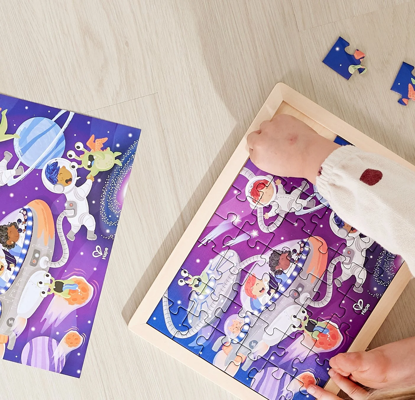 A person assembling a space-themed Hape jigsaw puzzle featuring astronauts and aliens on a purple background on a light-colored table.