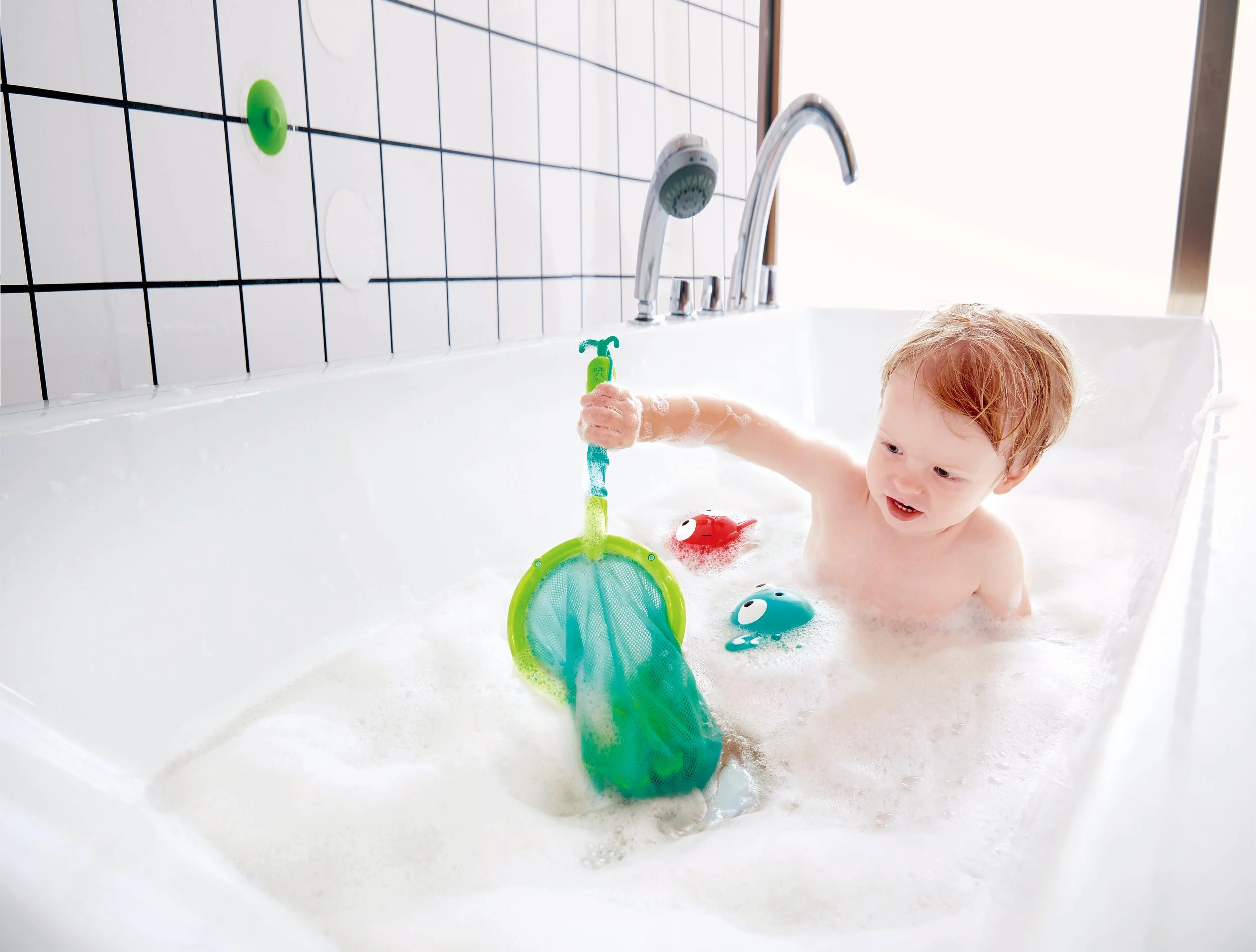 A young child playing with "double fun fish set" by Hape in a bubble-filled bathtub.
