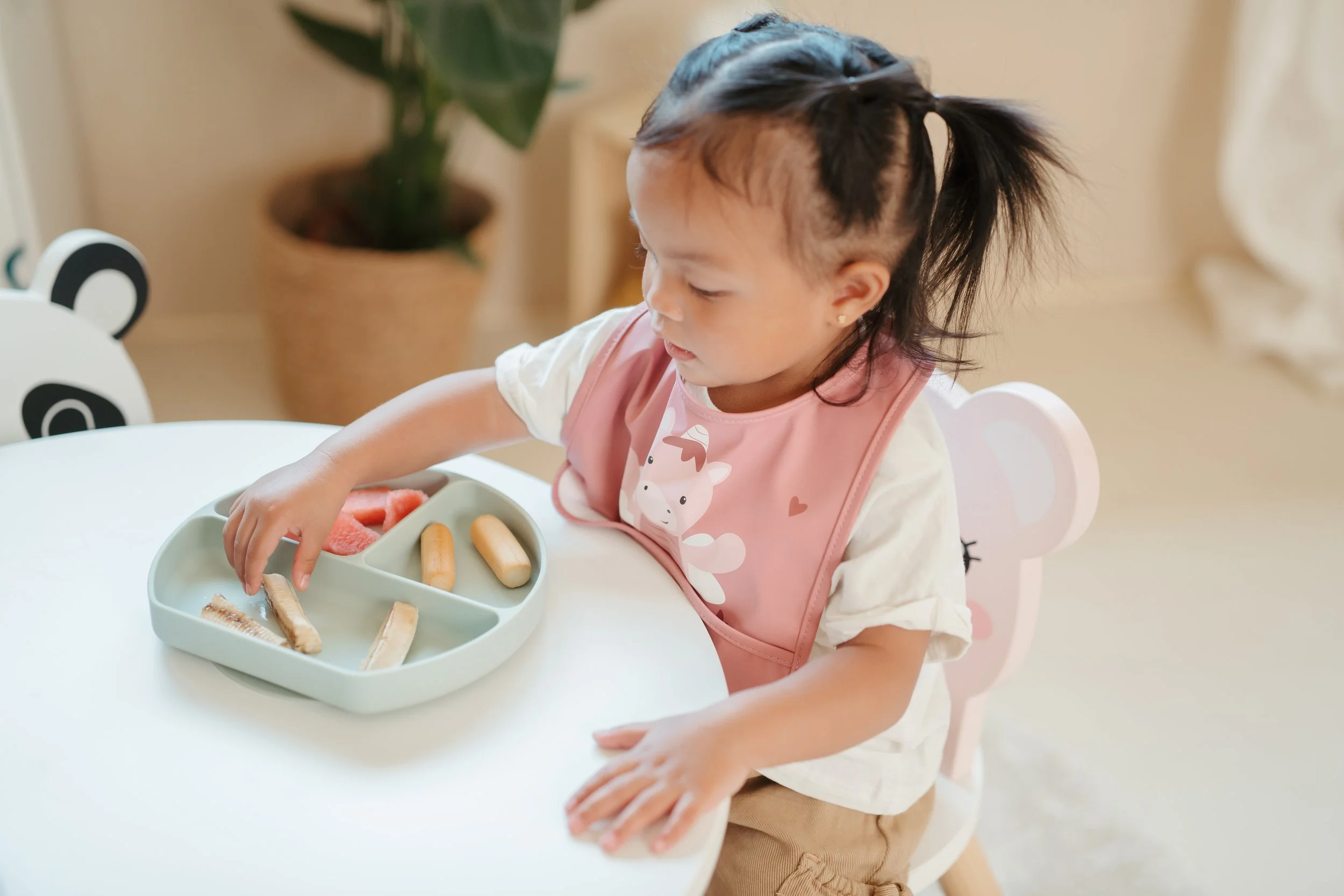 A young girl with a ponytail wearing a pink bib with a cow design, sitting at a white table, reaching for a snack from a divided tray with breadsticks and watermelon slices.