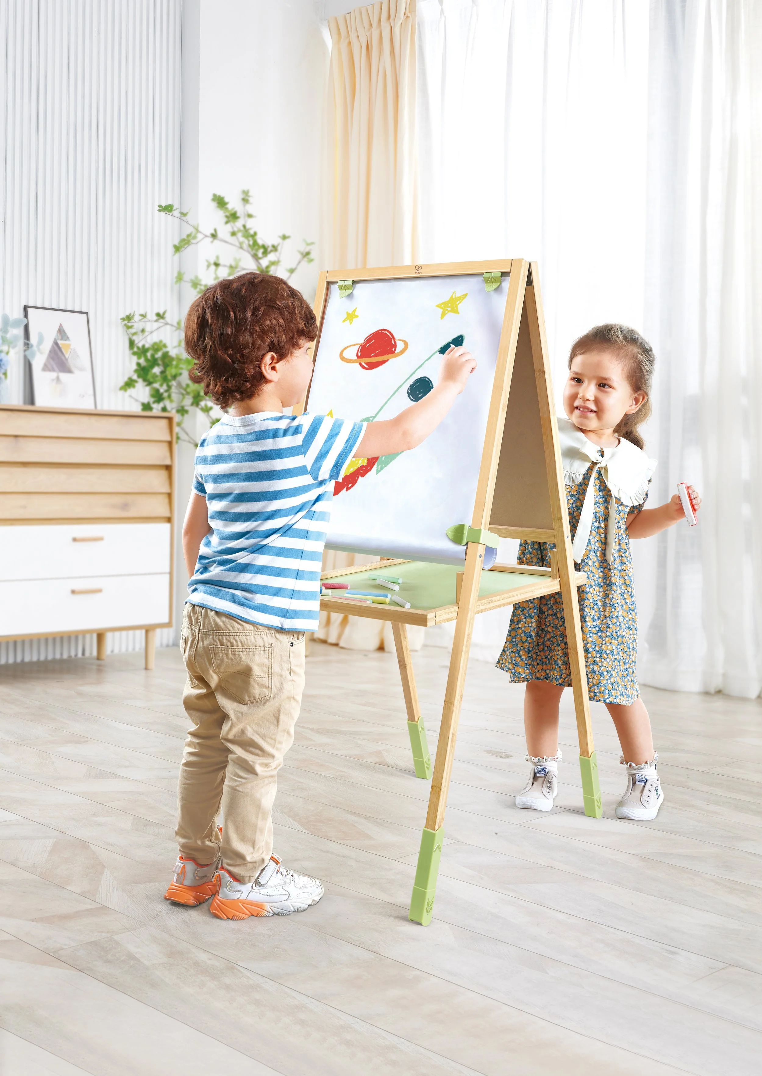 Two children drawing on the Hape whiteboard with space-themed illustrations, one boy and one girl, in a bright room with curtains and a plant.