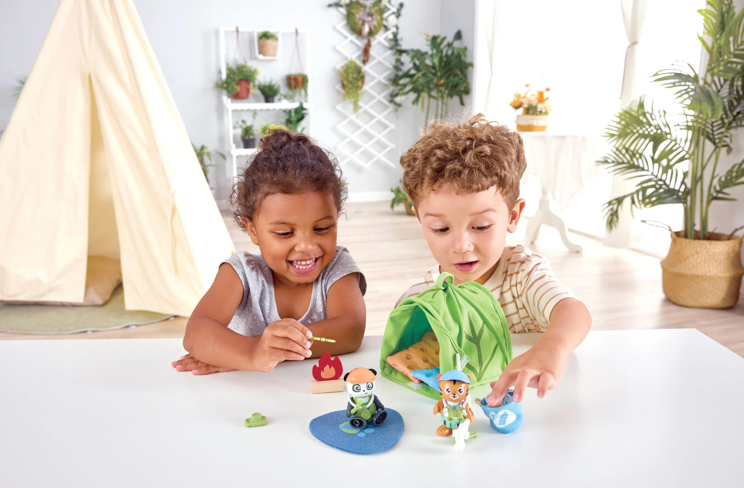 Two children, playing with Green Planet Explorers Camping playset by Hape at a white table inside a bright, decorated room.
