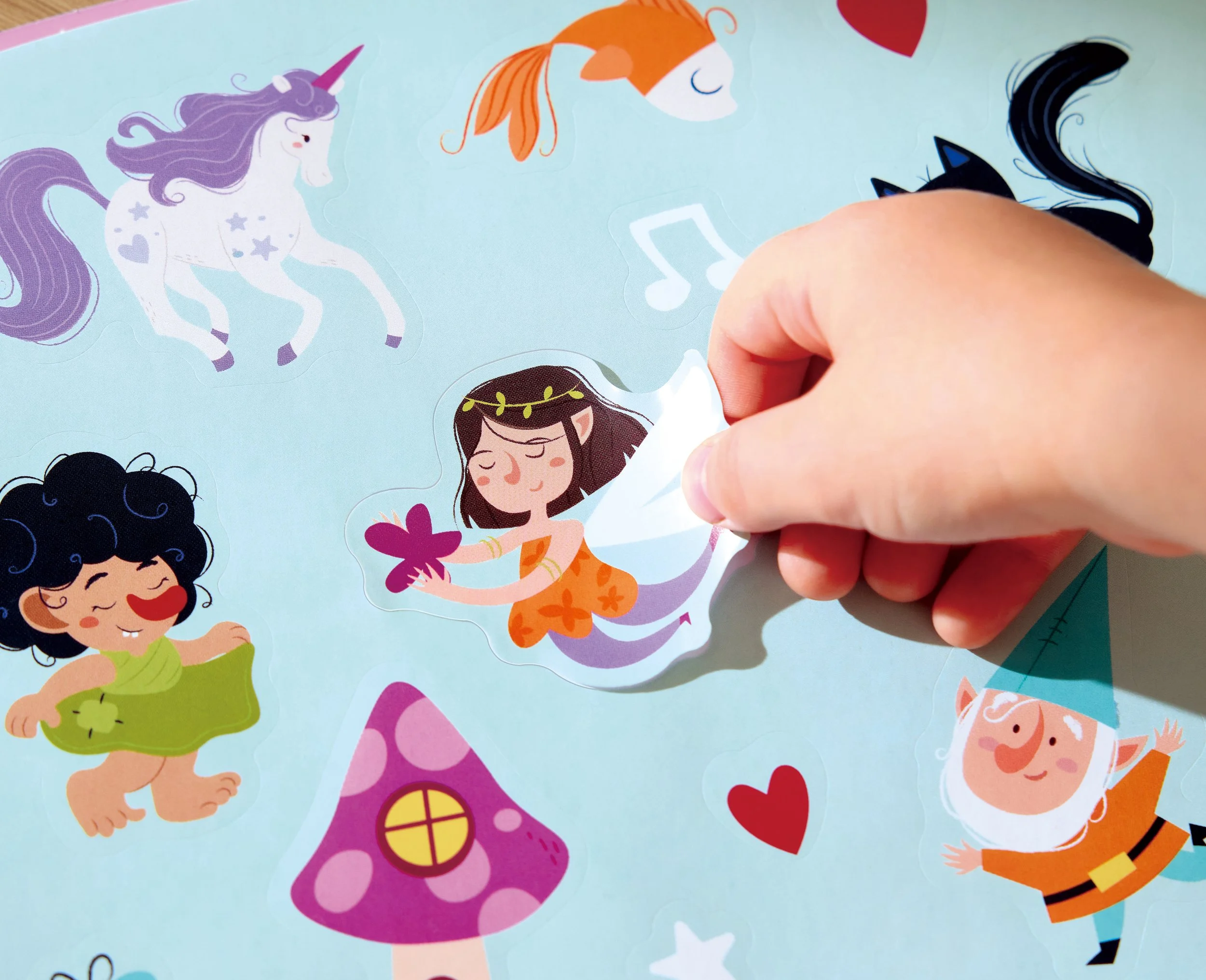 Child peeling off a sticker depicting a fairy with brown hair and an orange dress from a light blue background decorated with rainbows, hearts, mushrooms and unicorns.