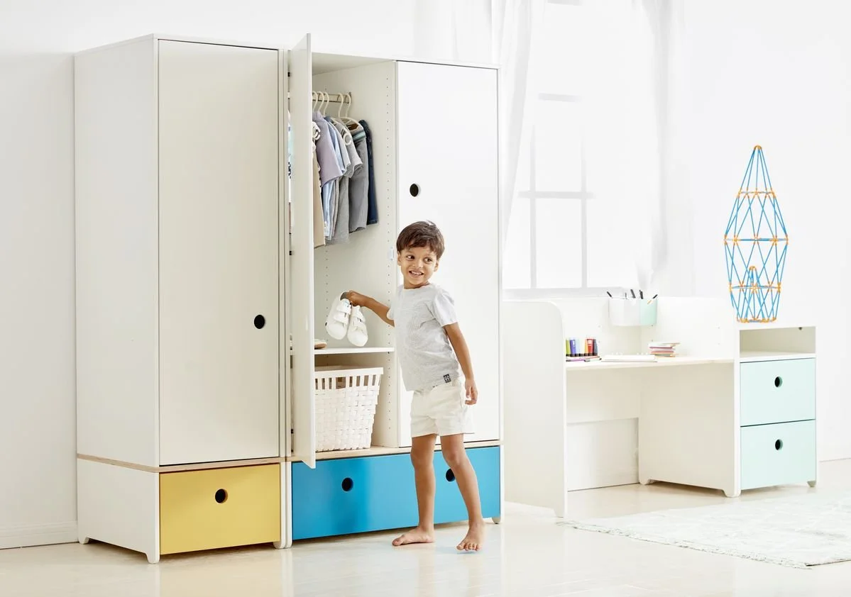 A young boy organizing shoes in a white Colorflex wardrobe with yellow and blue drawers in a bright, modern room with a white desk and decorative light fixture.