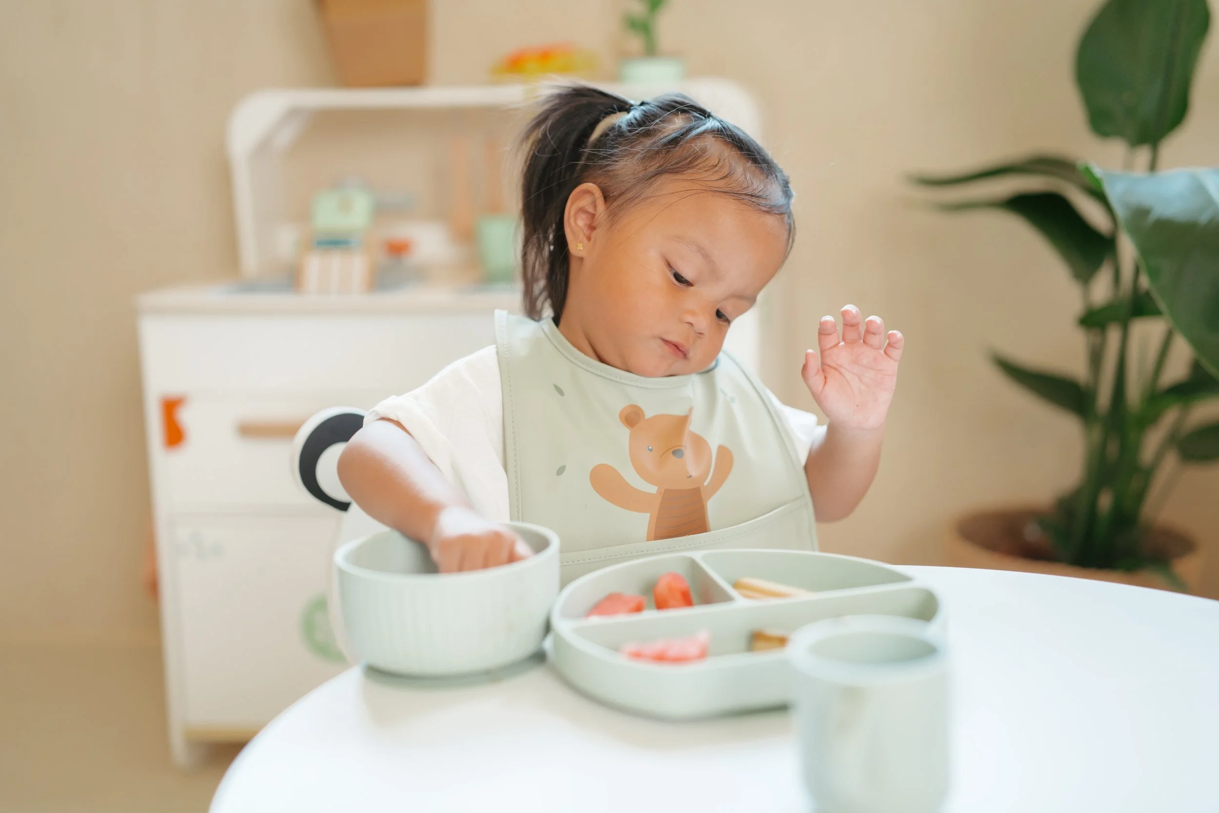 A young girl sitting at a table with a divided plate of food, reaching into a bowl, wearing a bib with a cartoon bear, and waving her hand.