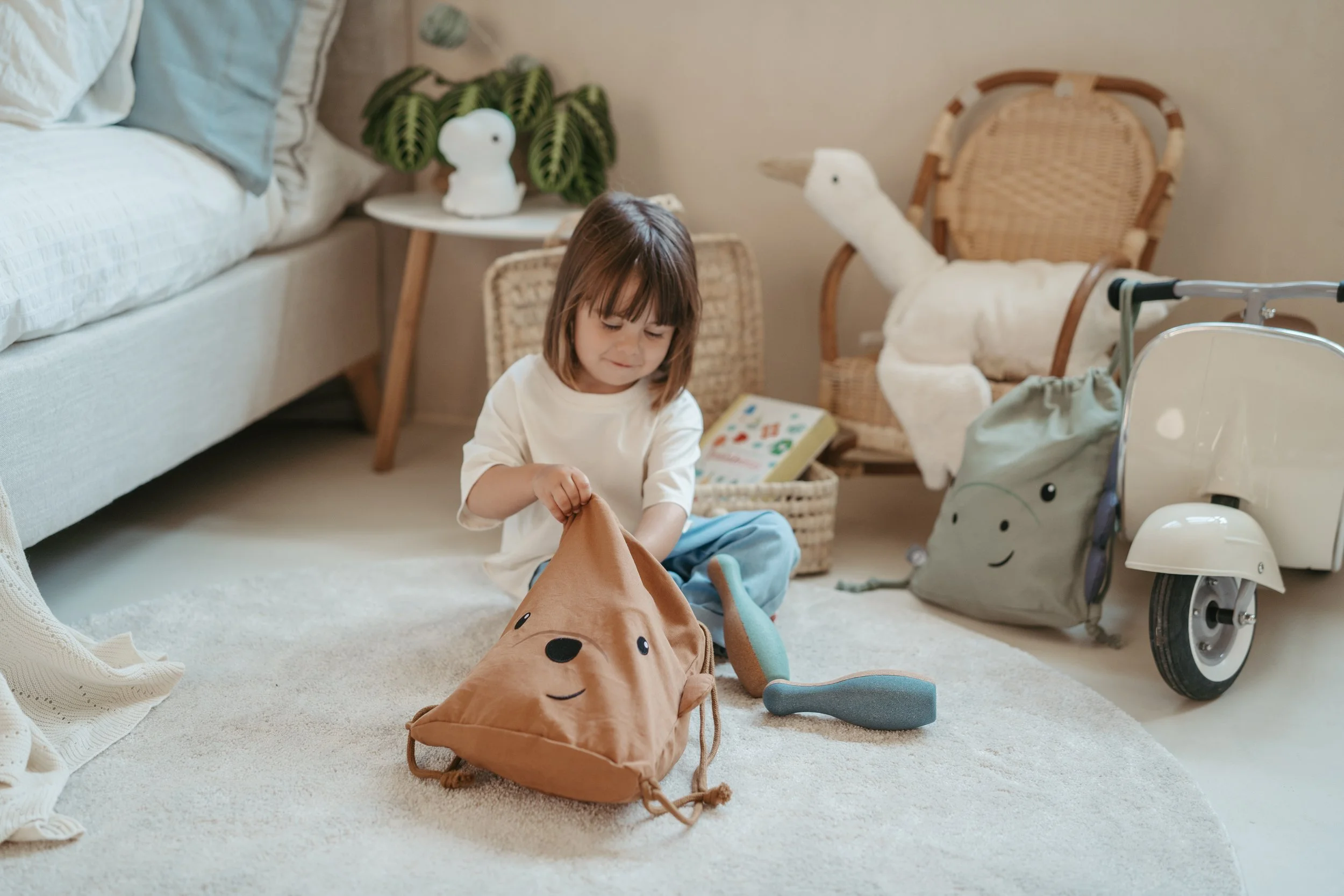 A young girl sitting on a rug, holding a plush bag with a bear face, surrounded by toys in a cozy living room with a sofa, wicker chairs, and playful stuffed animals.