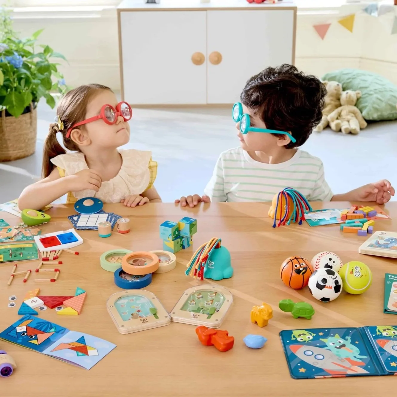 Two children sitting at a table wearing colorful glasses, playing with various toys, games, and puzzles in a bright, decorated room.