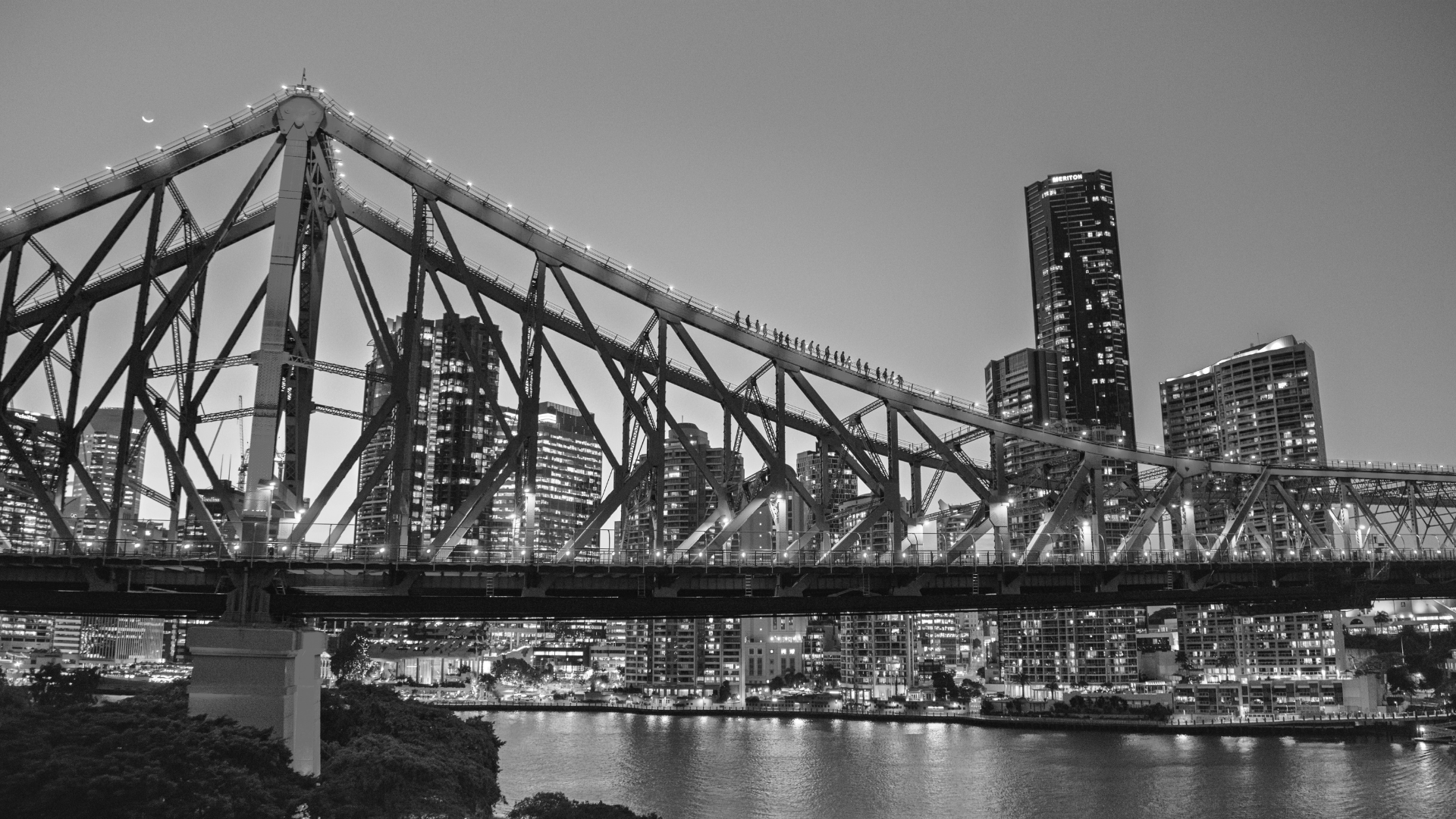 A black and white photo of a city skyline at night, featuring a large bridge with lights, tall skyscrapers, a river in the foreground, and a crescent moon in the sky.