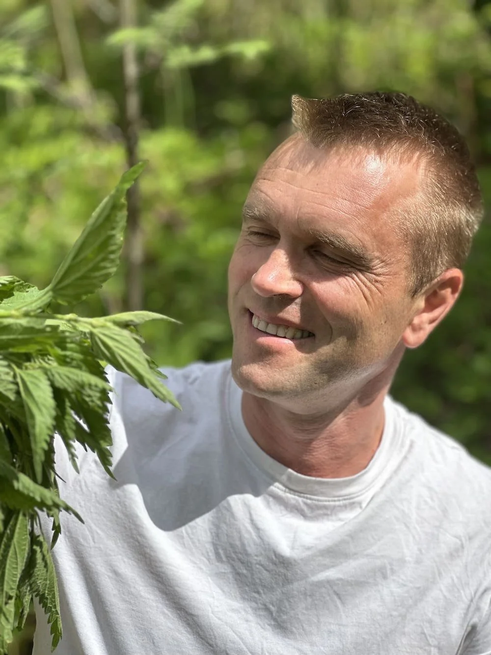 A smiling man with short brown hair smiling as he inspects a nettle plant outdoors in a green, wooded area.