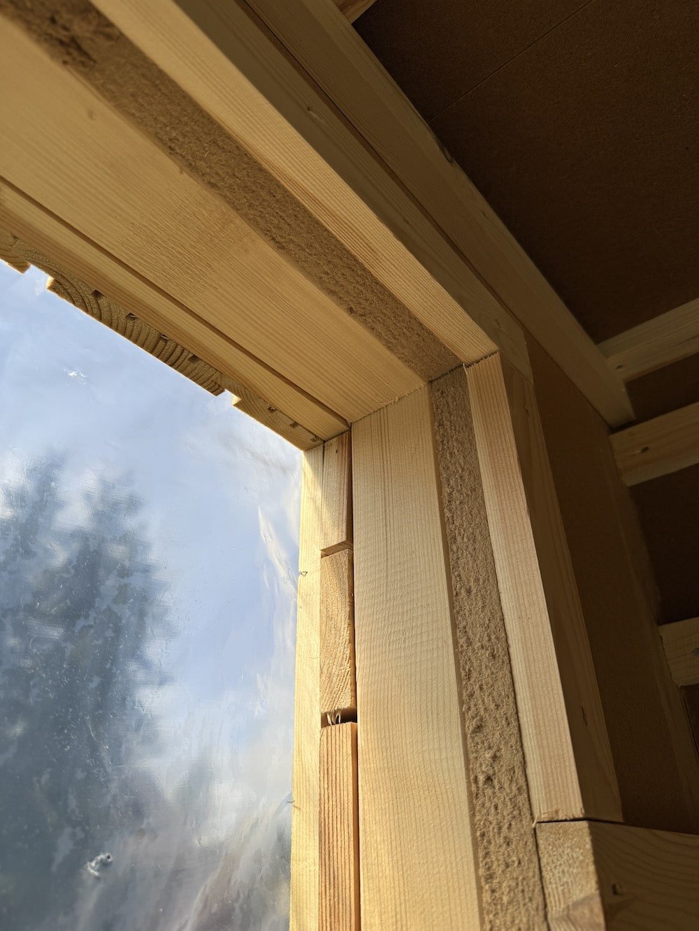 Close-up view of a window opening in a wooden structure under construction, showing framed wooden studs and blue sky with clouds outside.