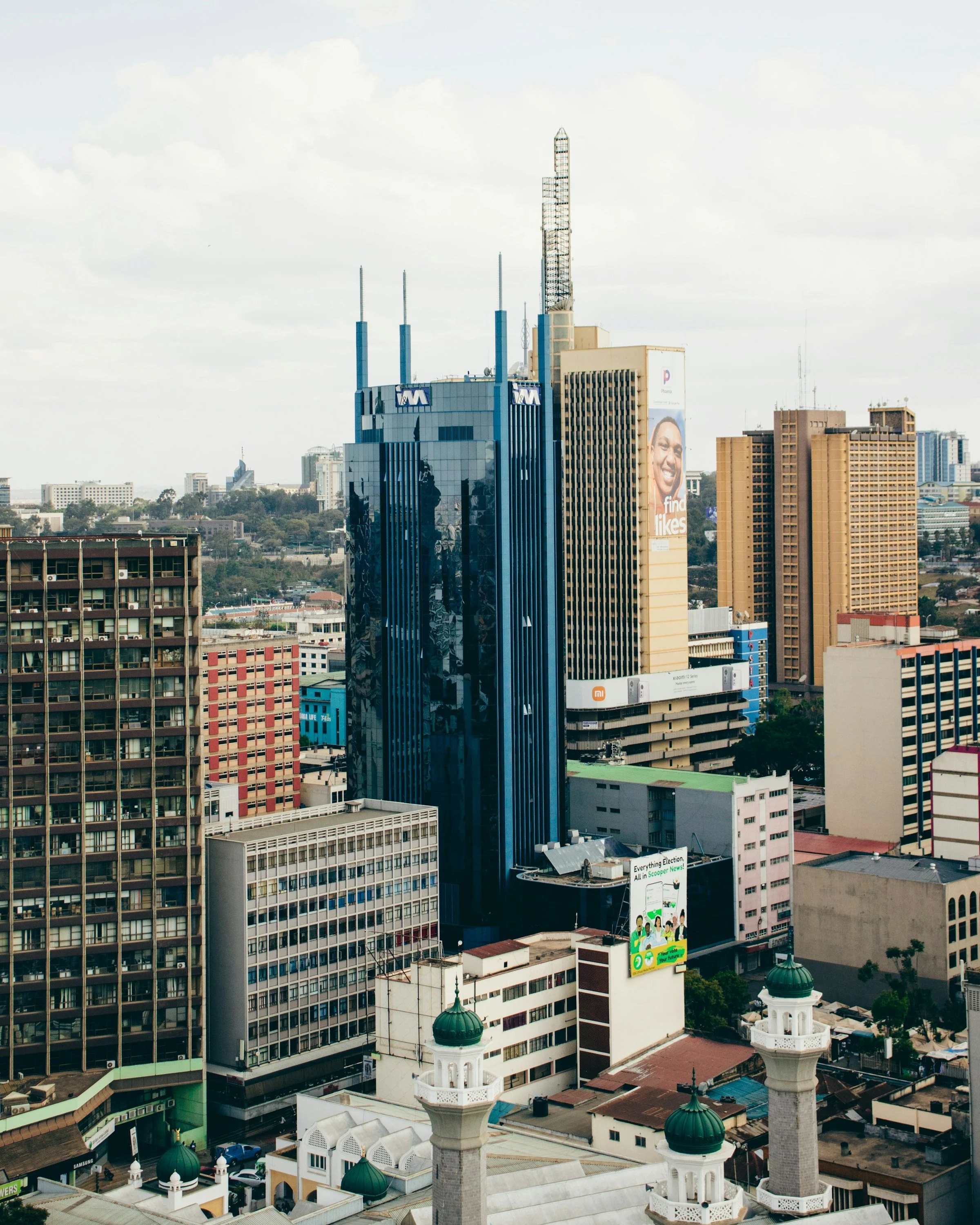 City skyline featuring tall buildings, including a prominent blue glass skyscraper with antennas, and four white minarets with green domes in the foreground.