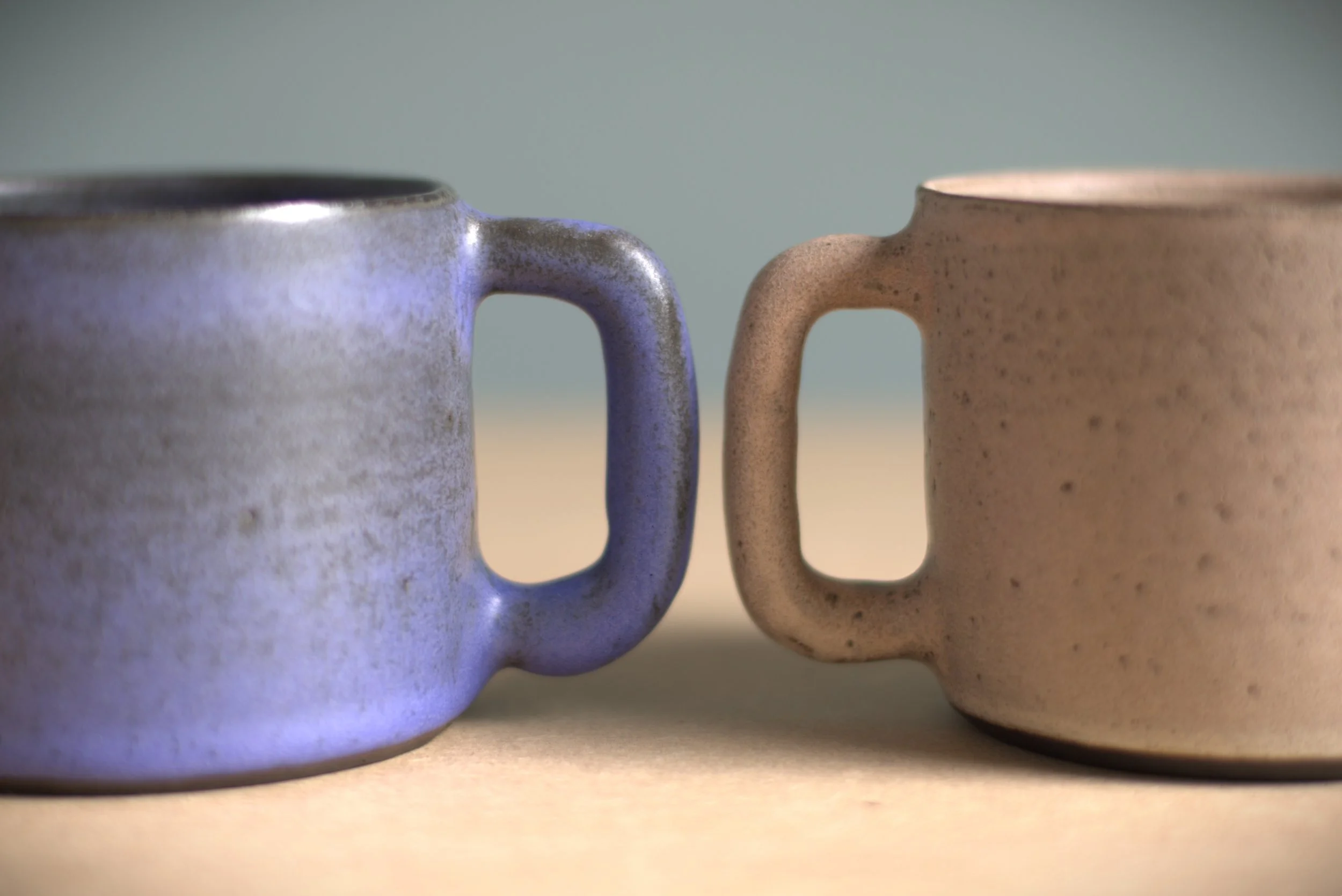 Close-up of two ceramic mugs facing each other, one blue and one beige, with similar square-shaped handles.