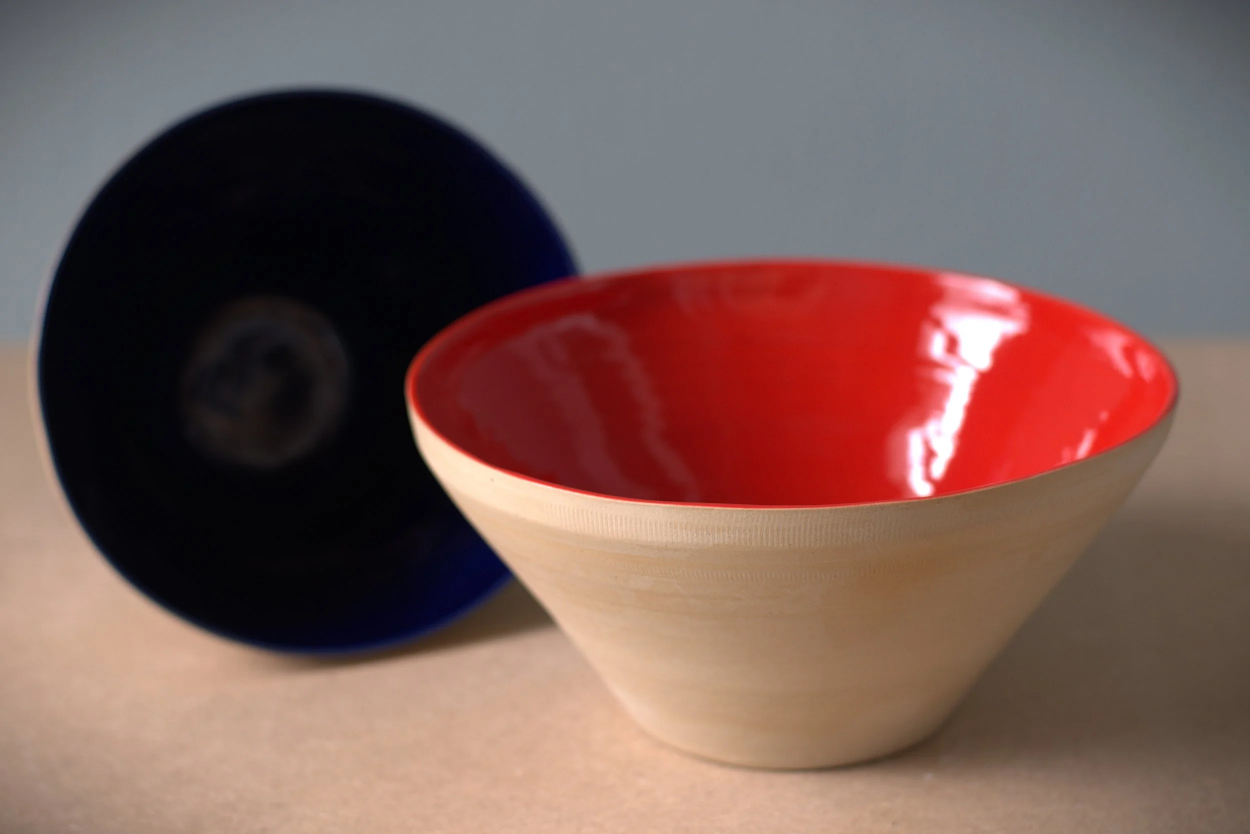 Two decorative bowls, one with a red shiny interior and the other with a dark interior, placed on a beige surface with a gray background.