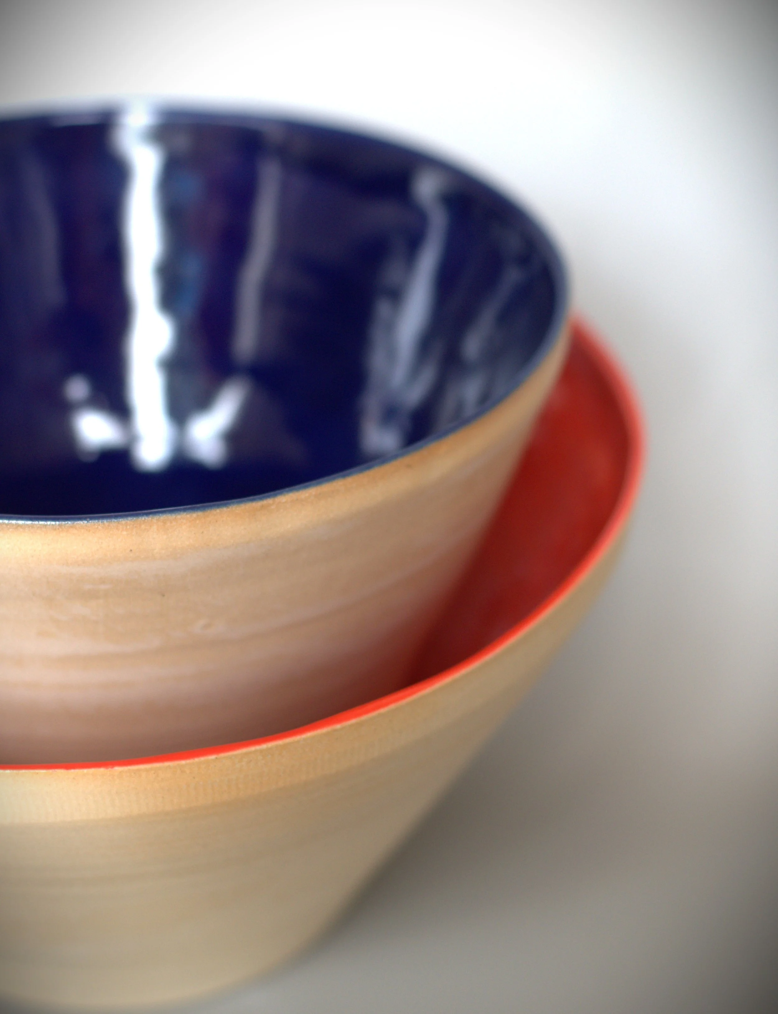 Close-up of two stacked ceramic bowls, one with a beige exterior and red interior, and the other with a deep blue interior, placed on a white surface.
