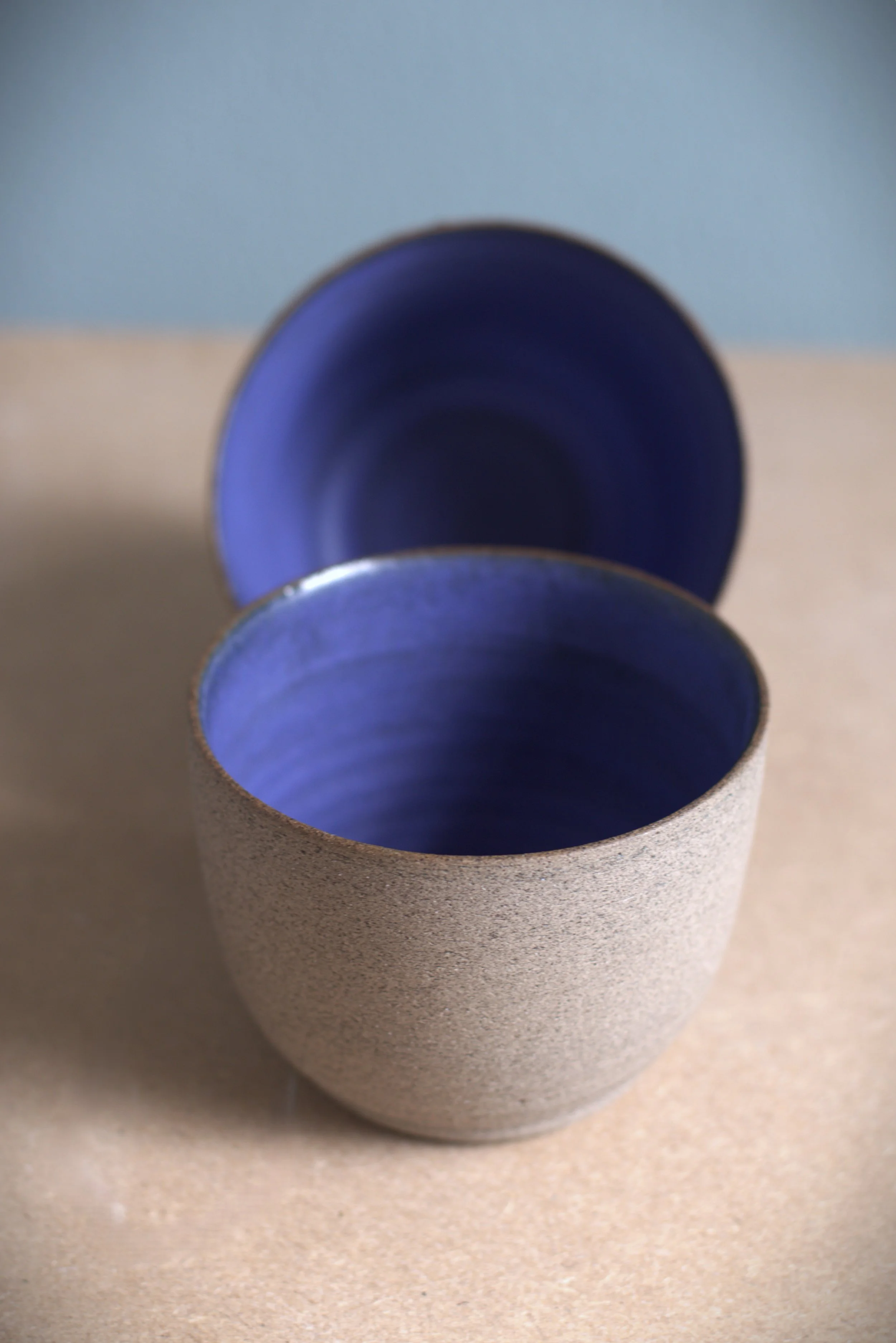 A textured beige ceramic mug with a deep blue interior, positioned in front of a matching blue cup on a beige surface with a blue background.