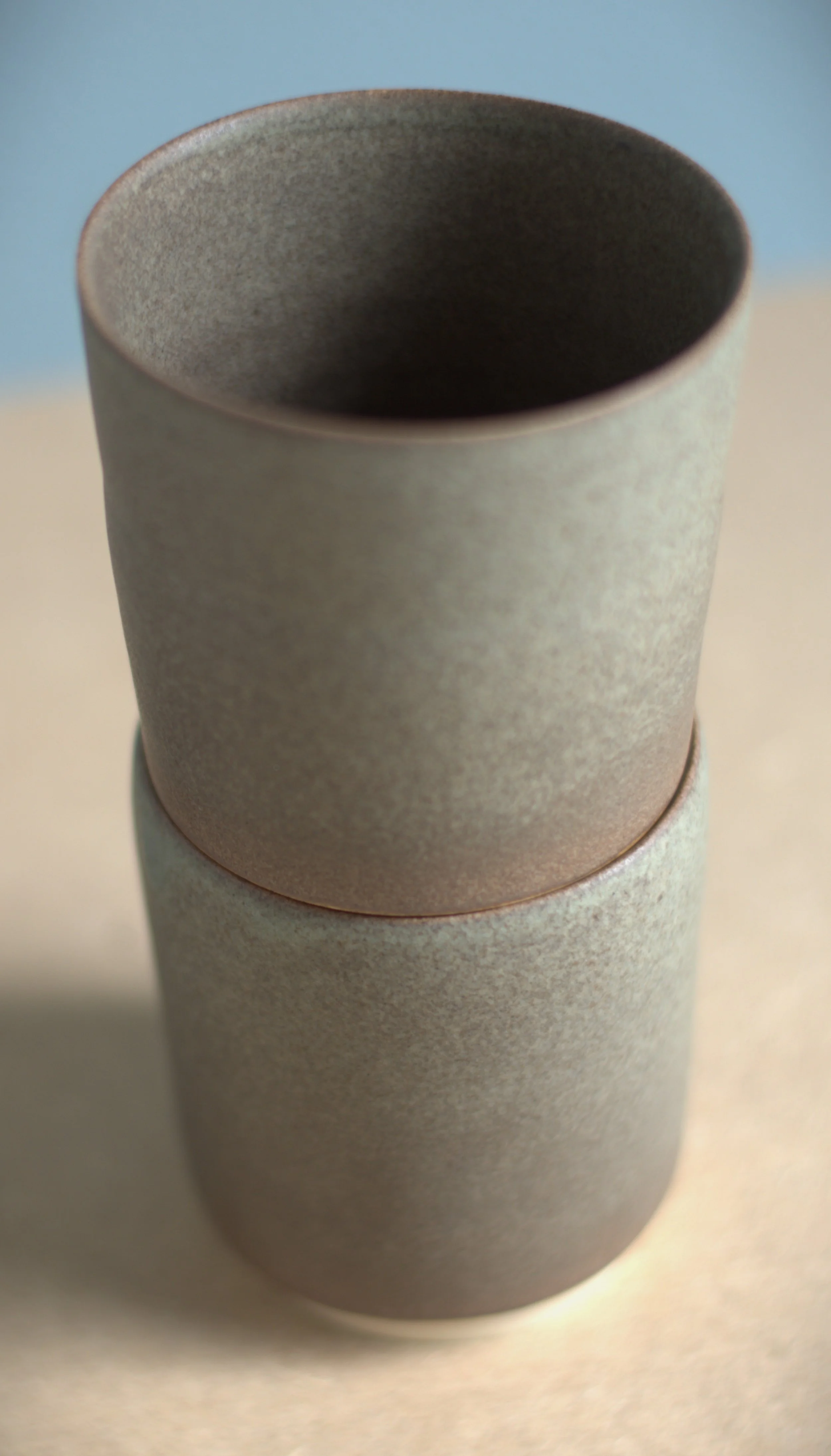 Two stacked matte beige ceramic cups viewed from above and to the side on a beige surface with a gradient blue background.