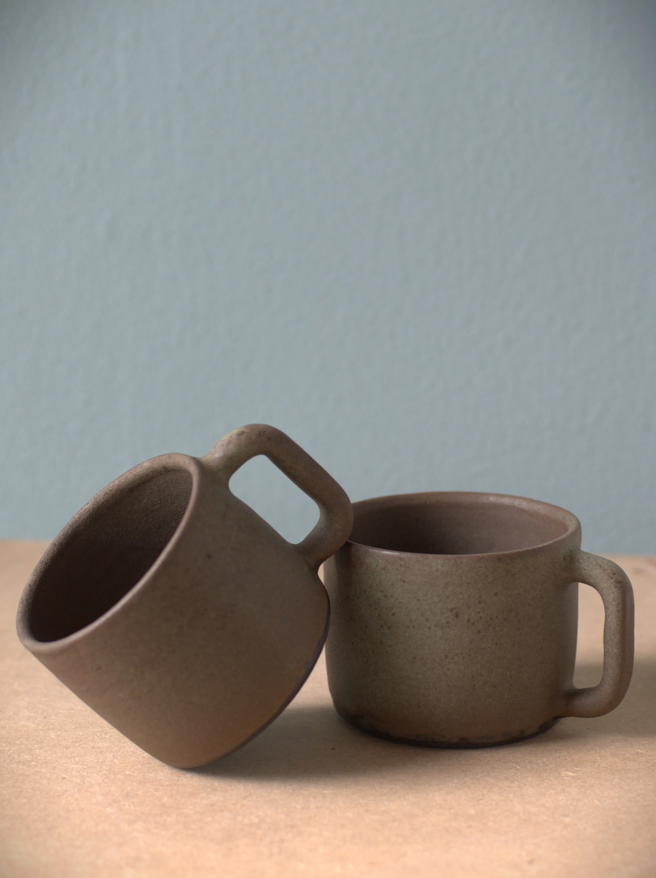 Two brown ceramic coffee mugs with handles placed on a light-colored surface against a plain background.