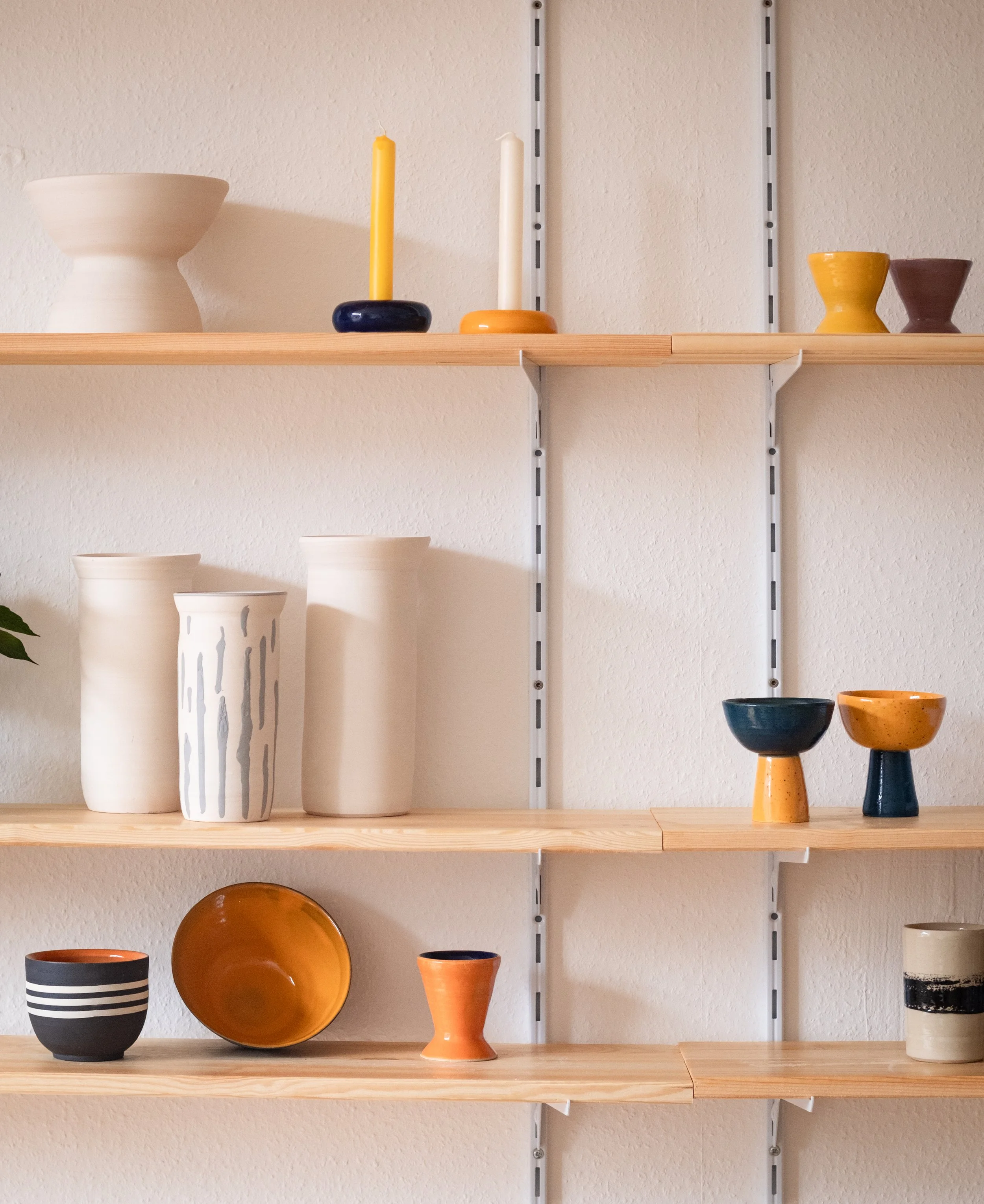 Wooden shelves with various minimalist ceramic and pottery vases, bowls, and candles against a beige wall.