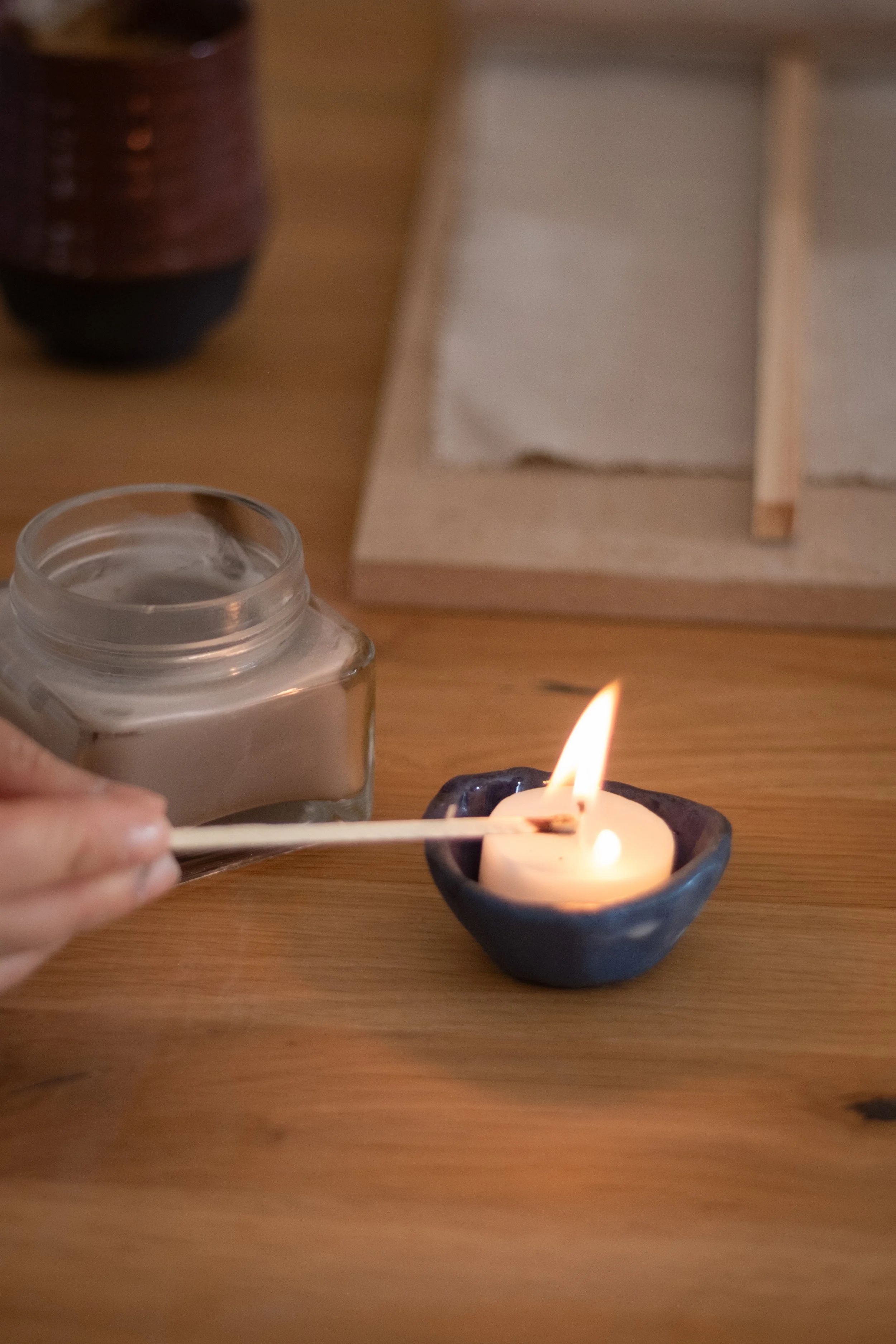 A person's hand is using a matchstick to light a small, lit candle in a black ceramic holder. There is a jar of candle wax nearby and a wooden tray with a cloth napkin in the background on a wooden surface.