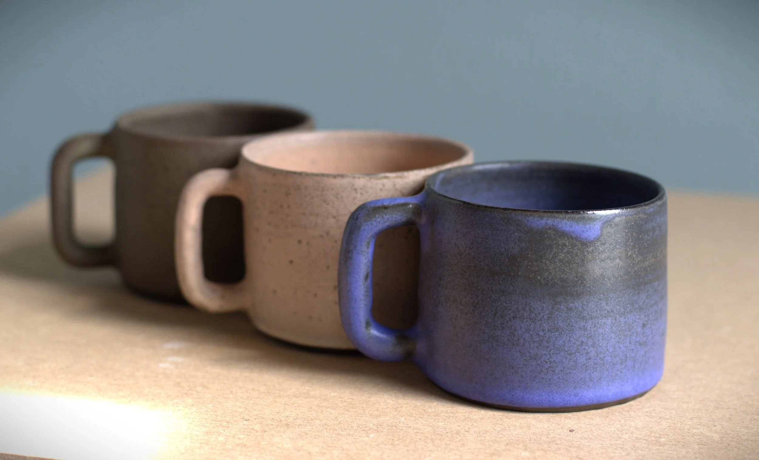 Three ceramic mugs in brown, beige, and blue, arranged in a diagonal line on a beige surface against a muted blue-green background.