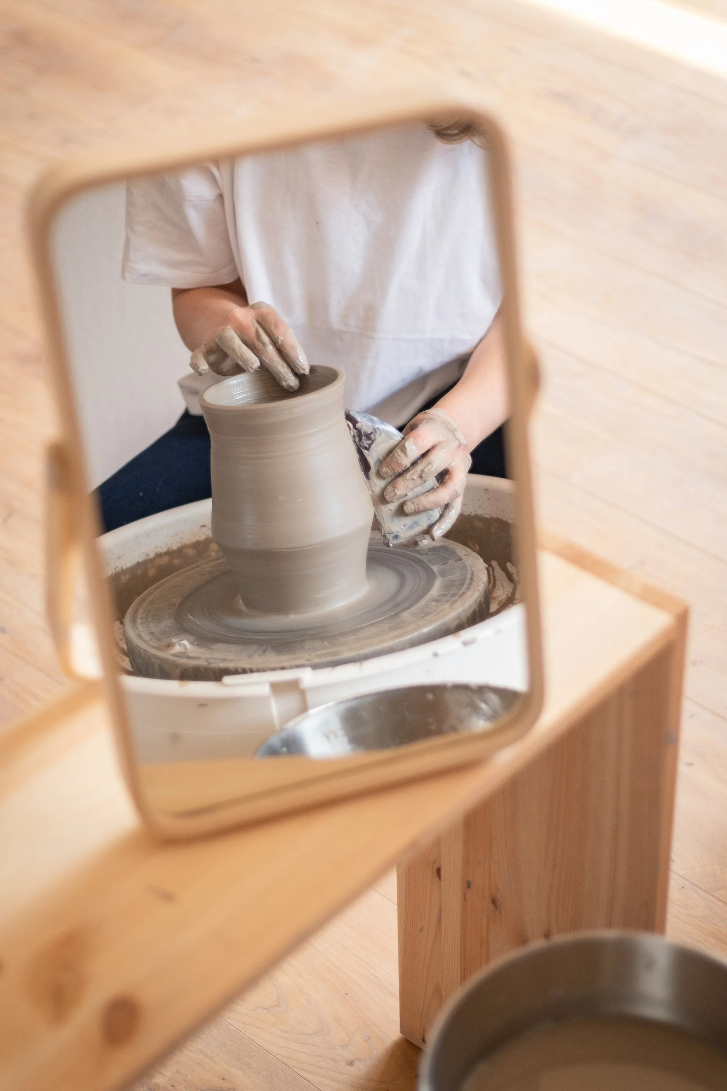An open studio participant works on the pottery wheel with a tool and throws a vase at open studio. They work in a fully equipt and peaceful place.