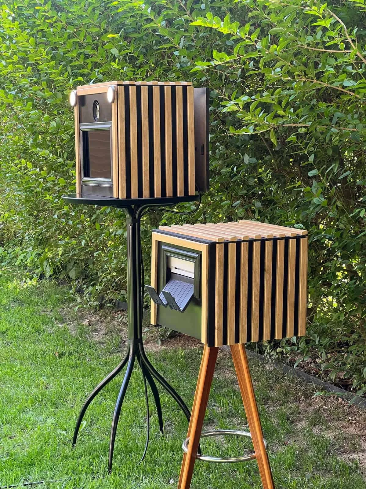 Deux cabines photobooth en bois avec des cadres en bois et en noir, positionnées en extérieur sur un support noir et un tréteau en bois, avec une haie de feuilles vertes en arrière-plan.