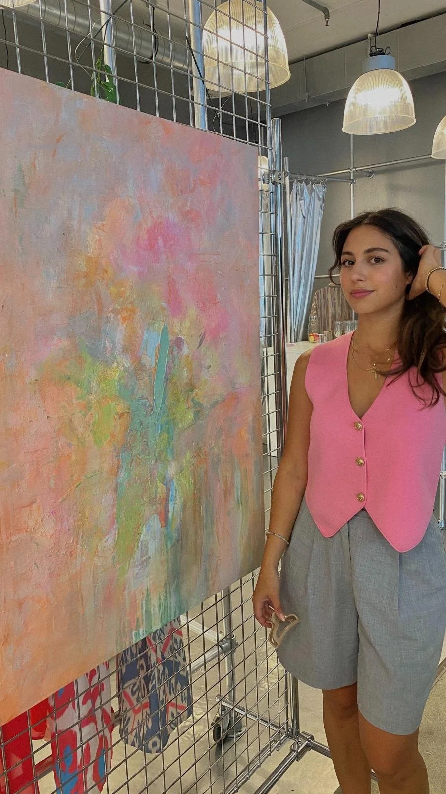 Junge Frau mit braunen Haaren, pinkem Weste und grauen Shorts steht neben einem abstrakten Gemälde an einer Metalldarstellung in einem Raum mit weißen Lampenschirmen