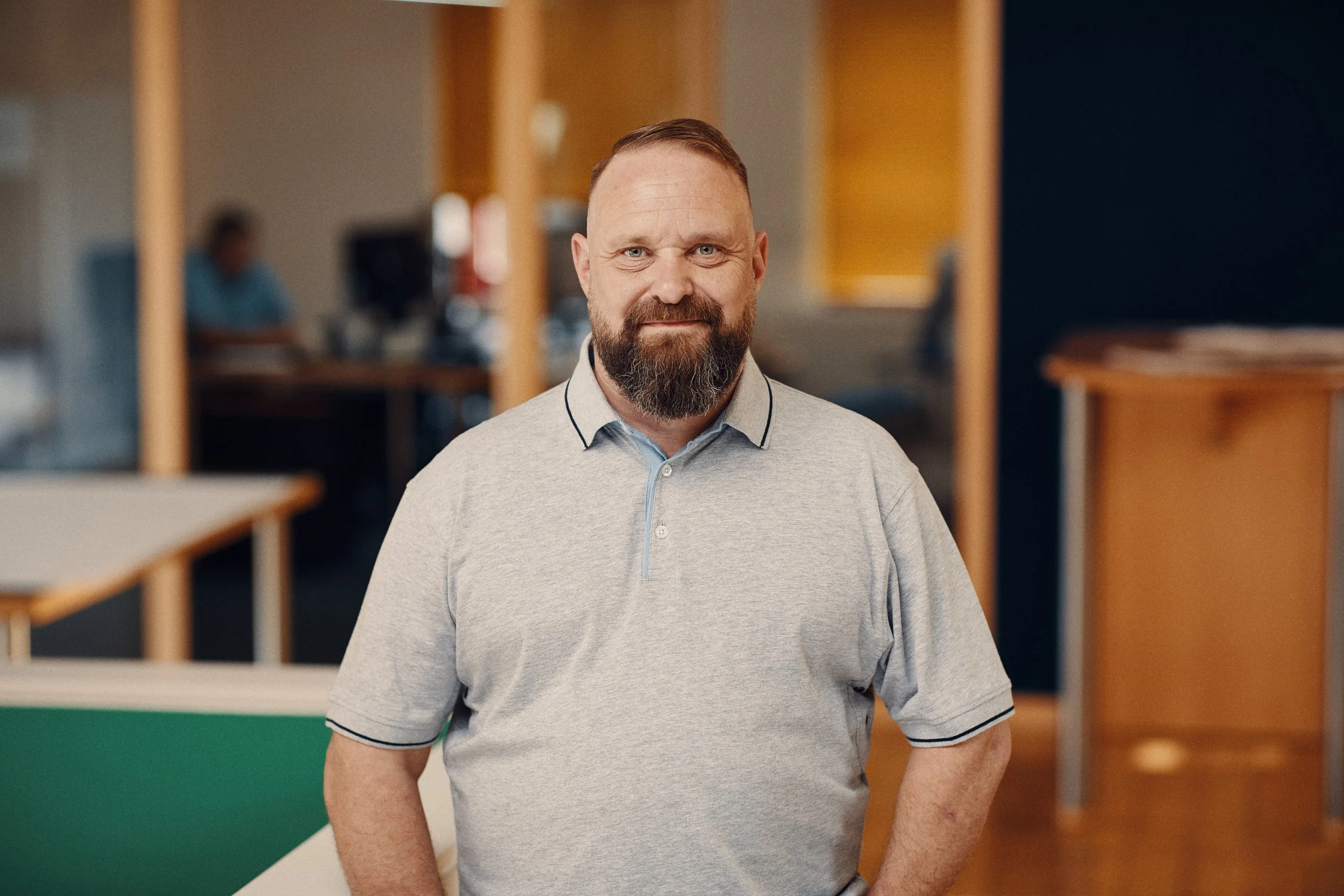 Ein Mann mit Bart und kurzen Haaren trägt ein graues Polohemd und steht in einem modernen Büro. Im Hintergrund sind andere Personen an Computern und Bürotische zu sehen.