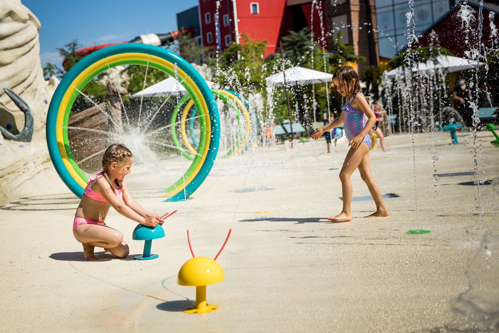 Zwei Kinder spielen im Wasser auf einem Spielplatz mit Wasserfontänen und Reifen im Freien bei sonnigem Wetter.