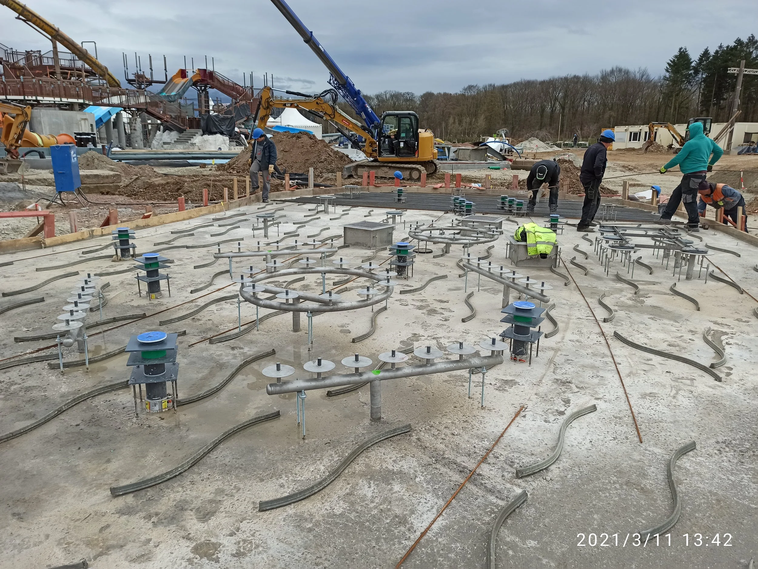 Baustelle mit Bauarbeitern, die an der Fundamentplatte eines Gebäudes arbeiten. Es sind verschiedene Rohre und Baugeräte auf der Baustelle zu sehen, sowie Bauhelme und -westen. Im Hintergrund sind Baumaschinen und Baumaterialien zu erkennen.