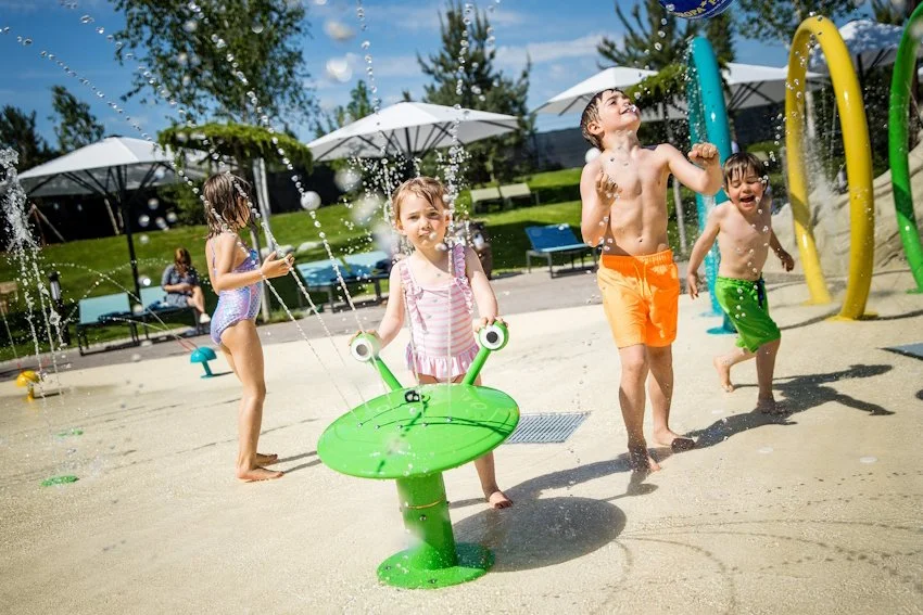 Children playing in the water park at water features under umbrellas on a sunny day.